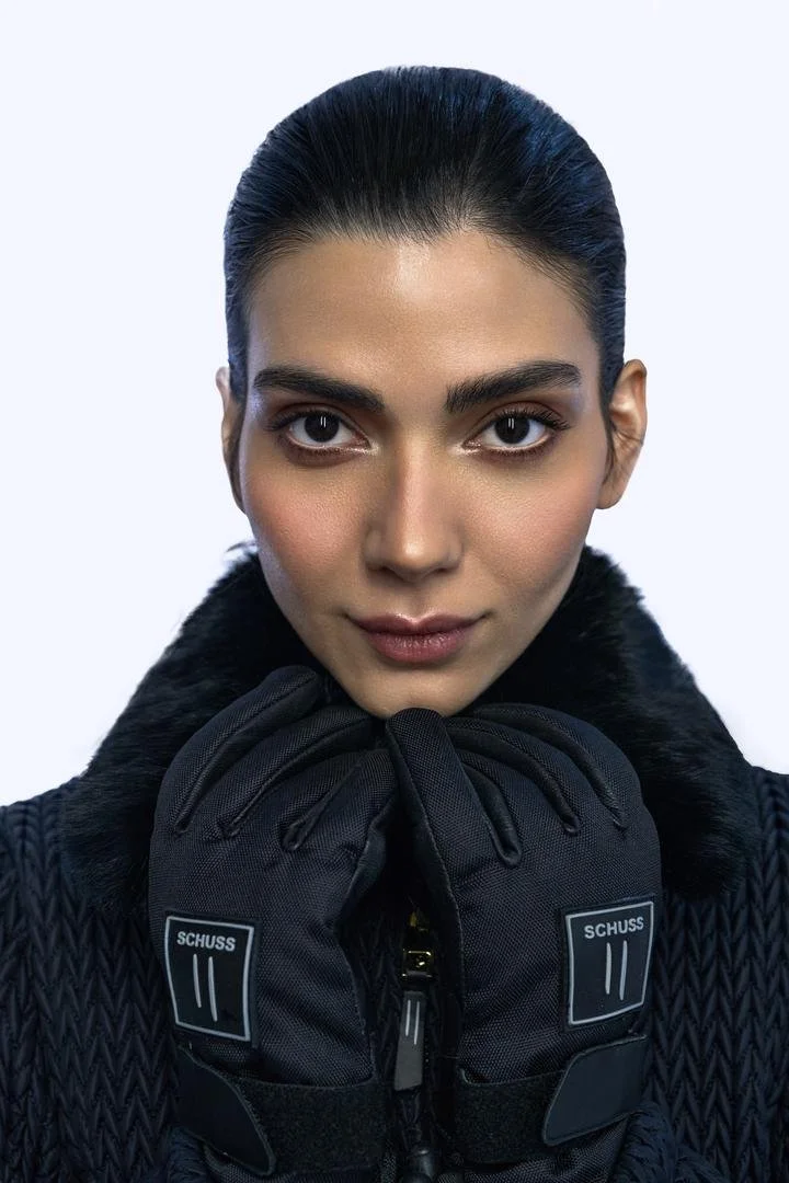 A woman with dark hair, slicked back, wearing a black textured jacket with a fur collar and black gloves, looking directly at the camera with a confident expression.