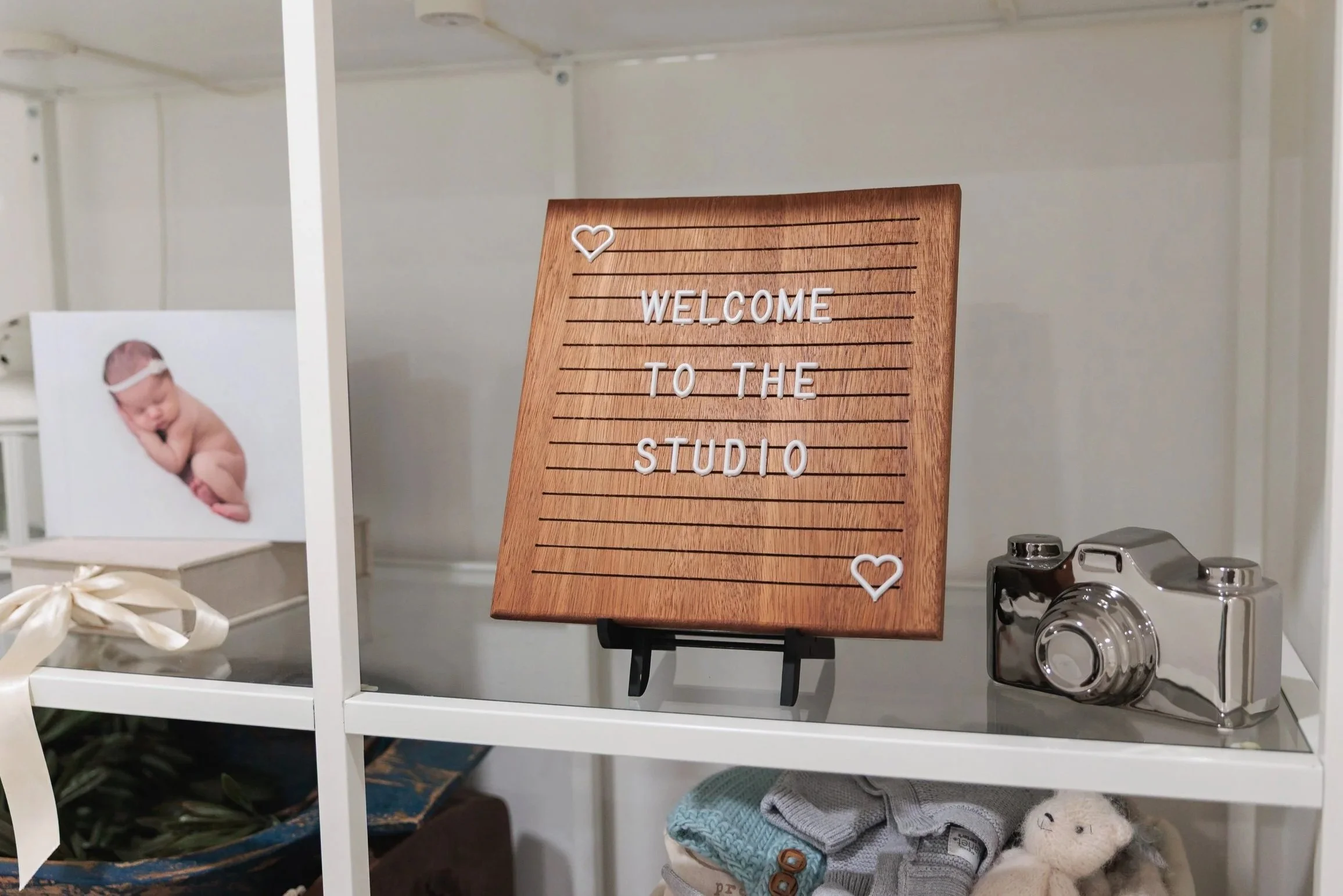 Wooden letter board with white plastic letters reading 'Welcome to the Studio', decorated with small white heart illustrations, on a black stand.