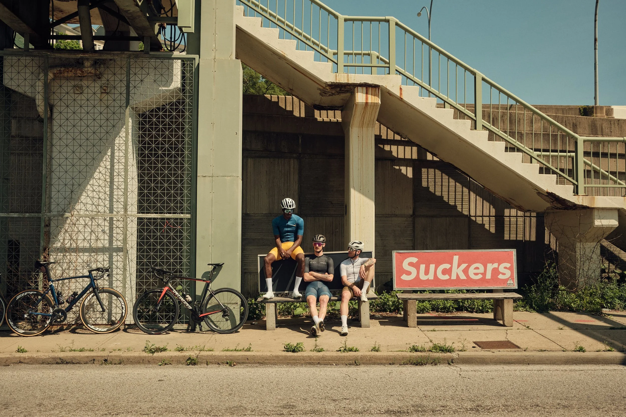 Four cyclists sitting on a bench under a bridge with two bicycles parked nearby, and a sign that reads 'Suckers'.