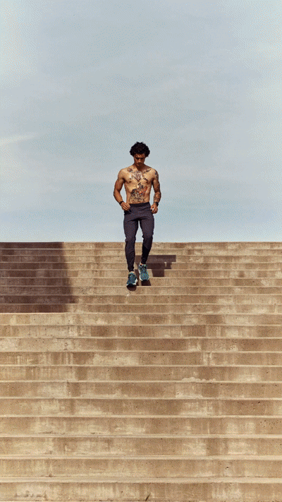 A man with curly hair, shirtless and covered in tattoos, jogging down a wide staircase outdoors under a cloudy sky.