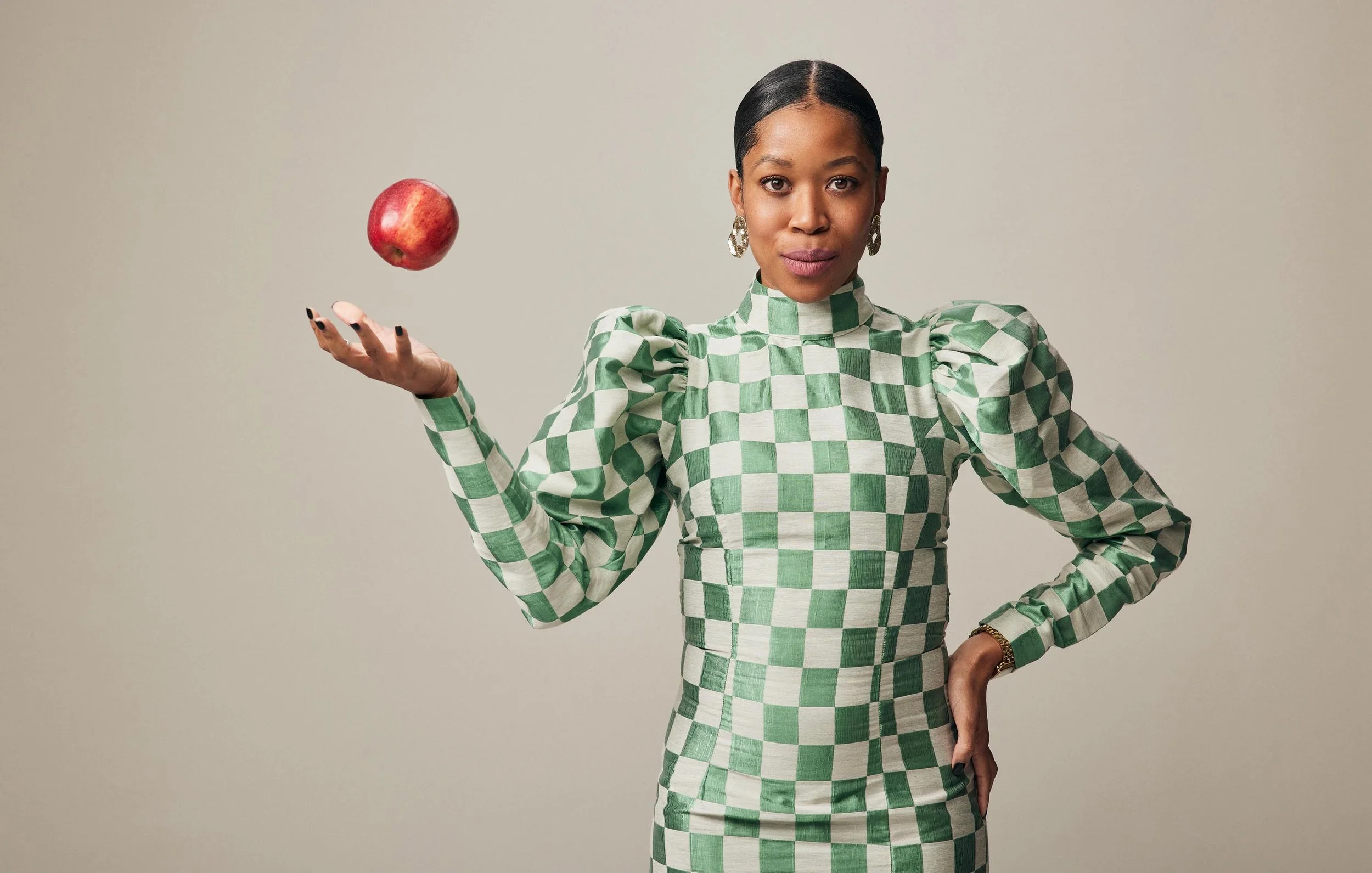 A woman with dark hair, wearing a green and white checkered dress with puffed sleeves, holding an apple in her left hand, standing against a plain background.