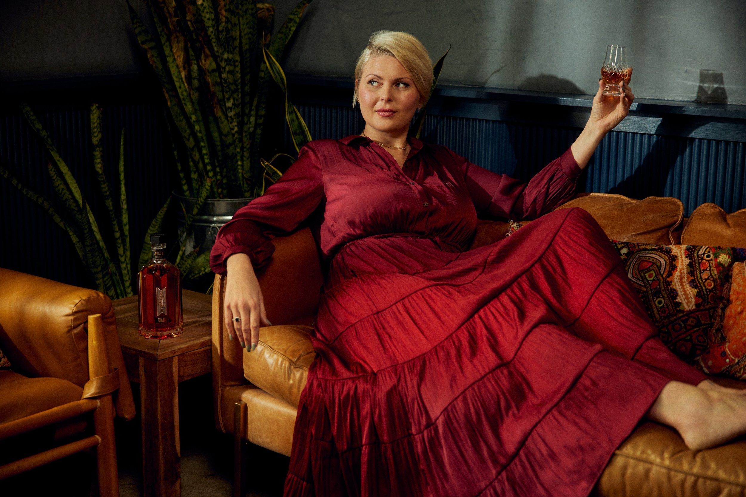 Marianne Eaves with blonde hair in a reddish dress sitting on a tan leather couch, holding a glass of what & Forbidden Bourbon captured for an advertisement by photographer Aaron Conway aaconn.
