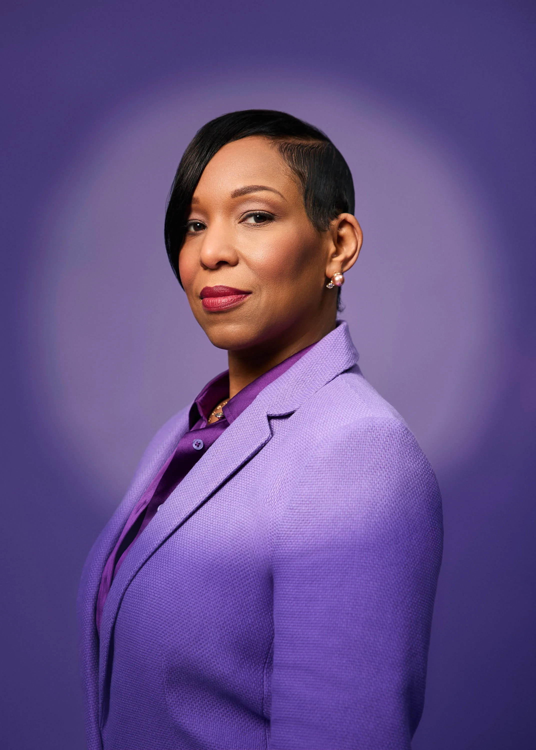 A professional woman with short black hair in a stylish side-swept haircut, wearing a purple blazer and matching shirt, against a purple background.