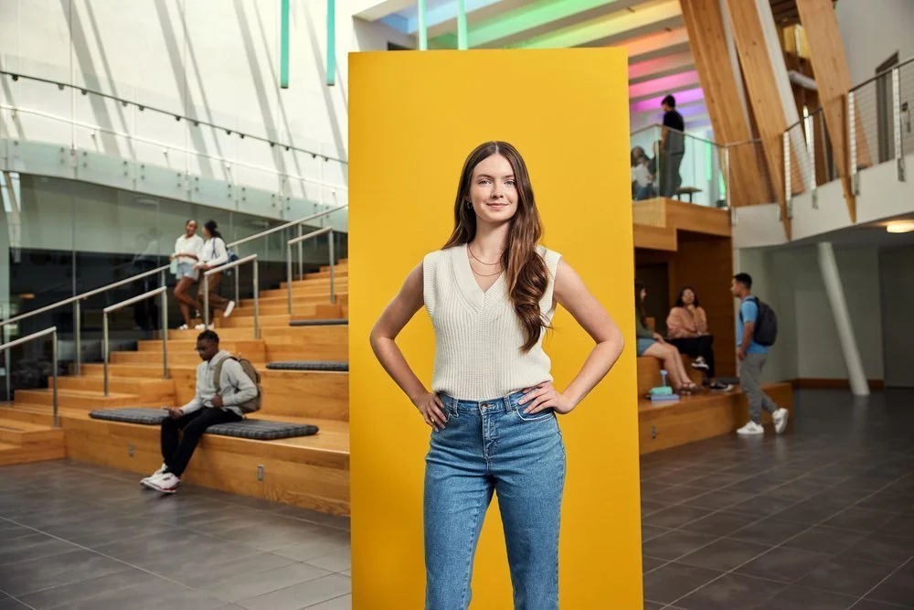 Young woman with long brown hair standing confidently in front of yellow backdrop in a modern, multi-level indoor space with wooden and glass design, people sitting and walking around.