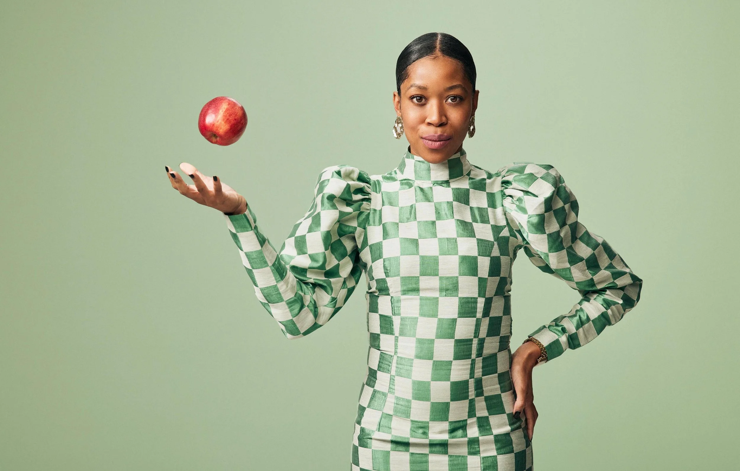 Photographer Aaron Conway captures. Krystal Howard in a green and white checkered dress standing against a solid green background, holding an apple in her raised right hand.