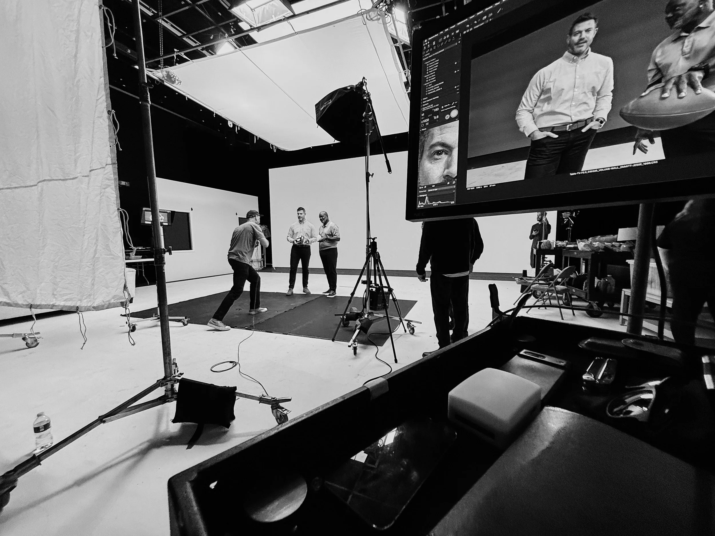 Scene inside a photography studio with crew members preparing for a photoshoot. Two men are posing for the camera while a crew member adjusts lighting. A large monitor displays a close-up of one model's face. Equipment like tripods, lights, and a workstation are visible around the studio.