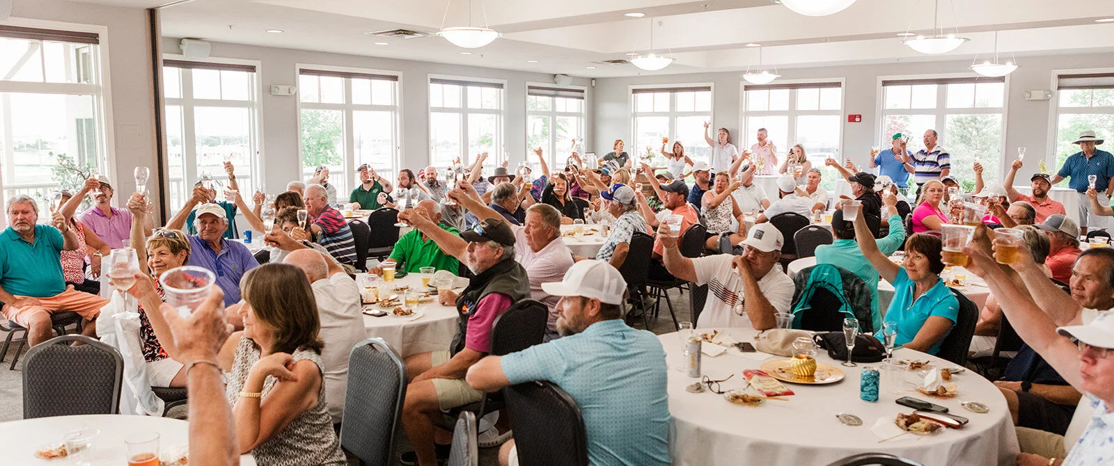 Large group of people gathered in a bright banquet hall, sitting at round tables, raising glasses for a toast during a celebration.