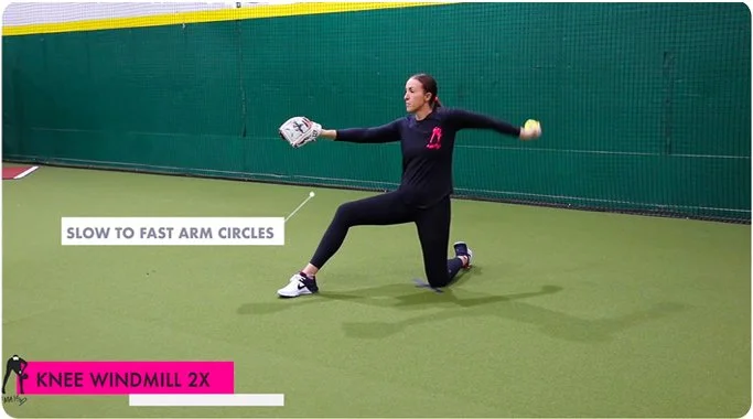 A person kneeling on one knee on an indoor turf field, wearing athletic clothing and holding a softball. They are demonstrating a pitch with the caption "Slow to Fast Arm Circles" and "Knee Windmill 2X" displayed on the image.