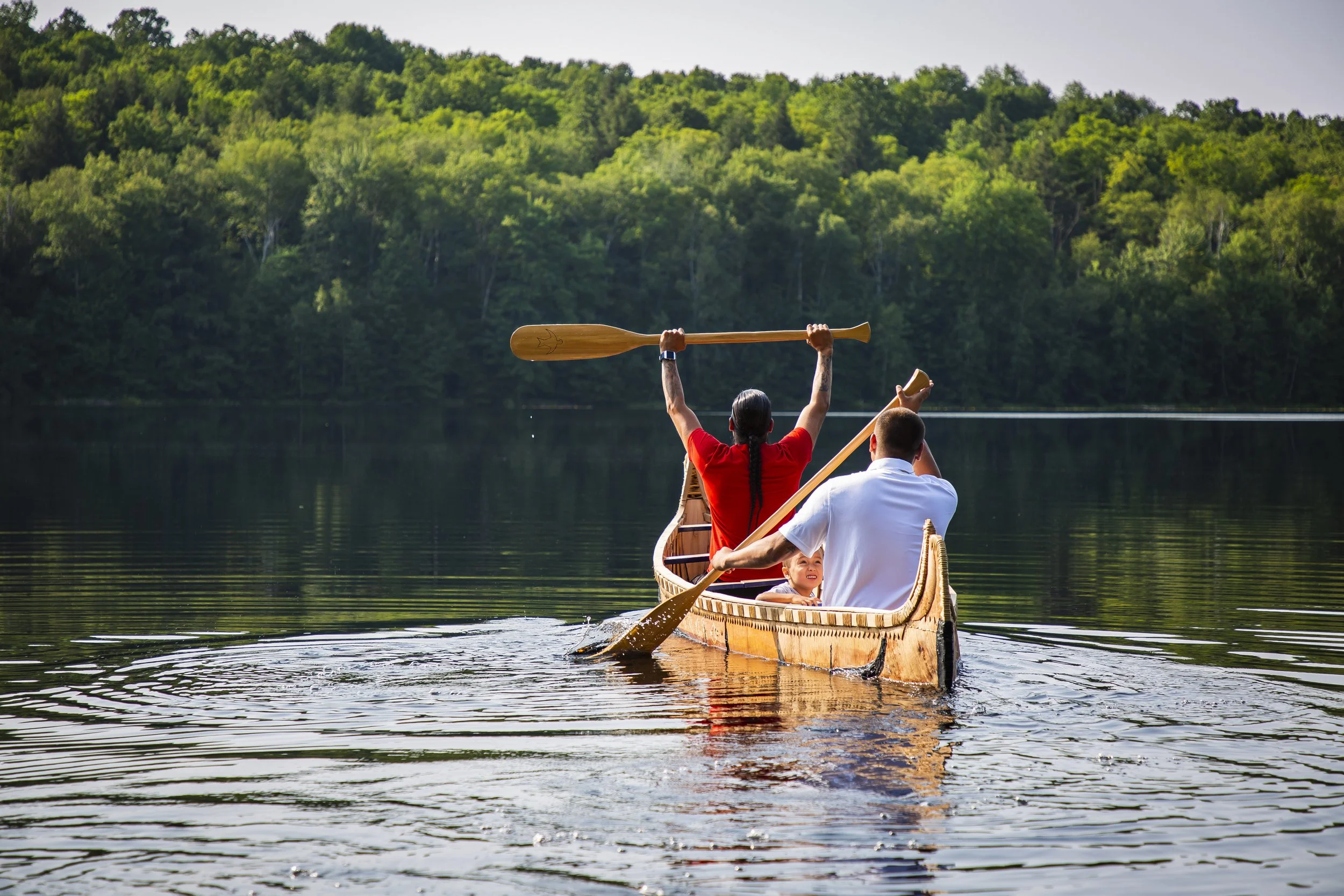 A group of people in a wooden canoe on a river, surrounded by green trees and hills, with one person holding a paddle raised in the air.
