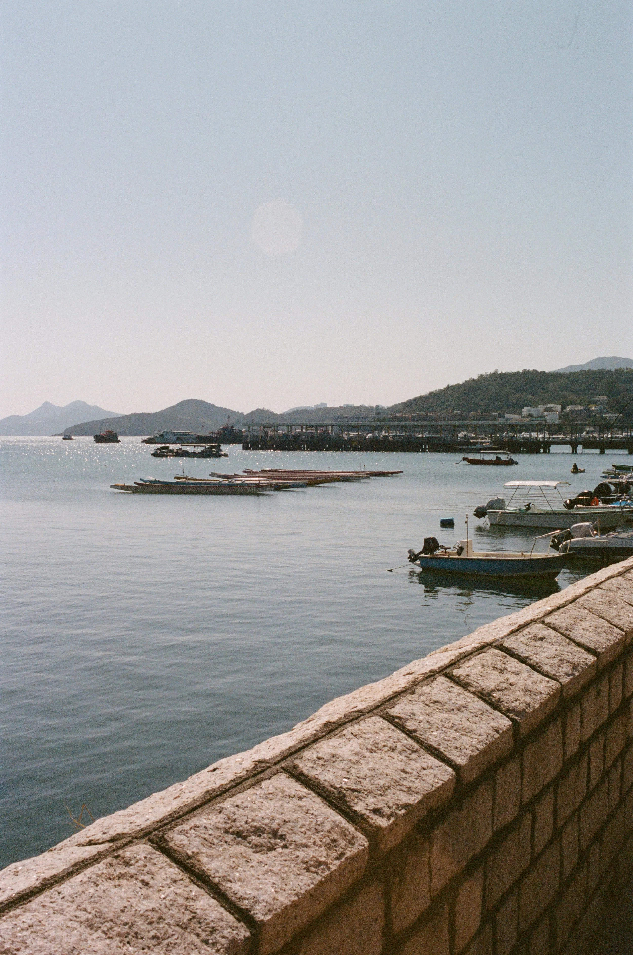 A peaceful harbor with small boats moored near a stone pier, distant hills in the background, and a clear sky.
