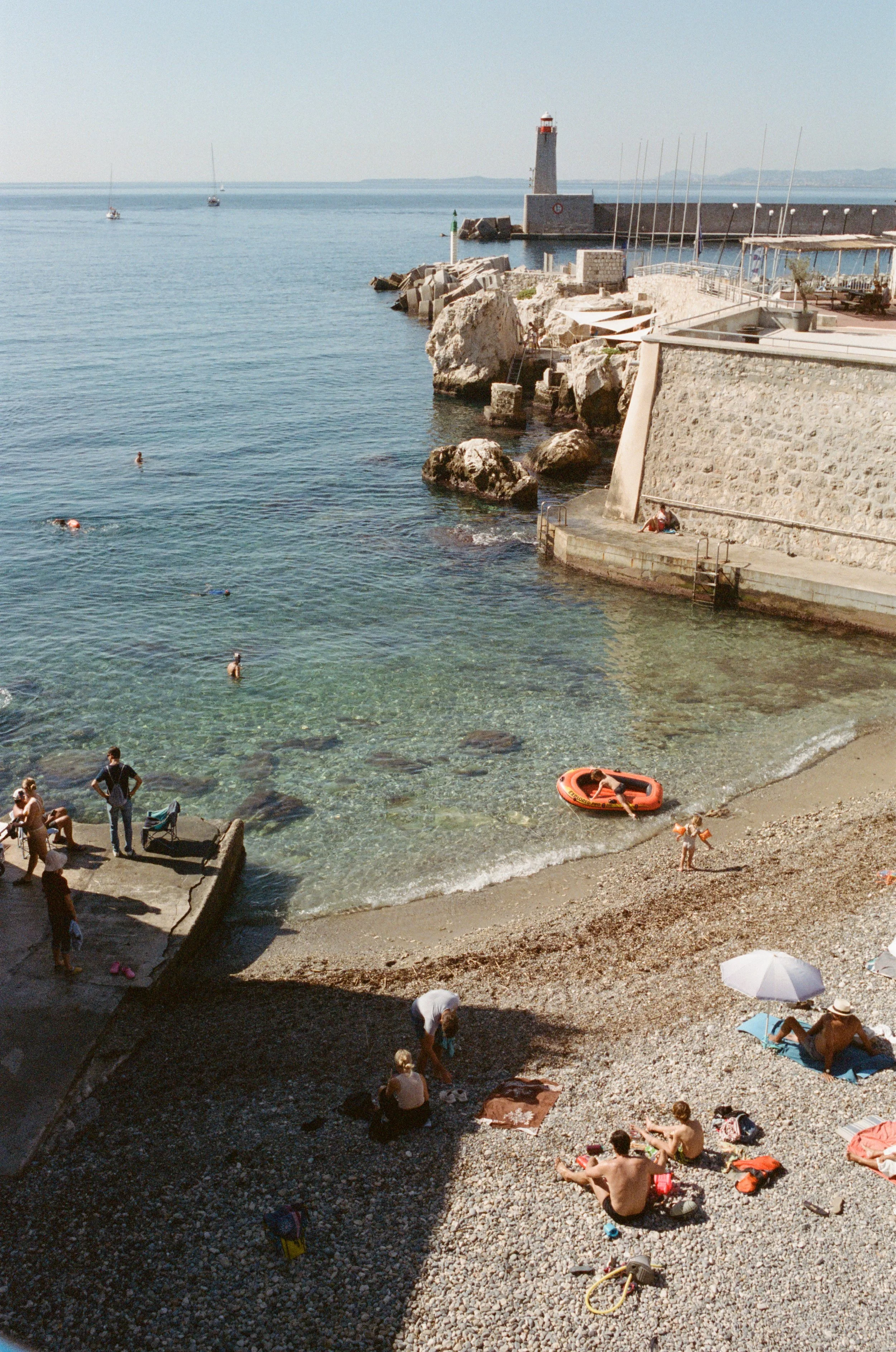 People swimming and relaxing on a pebbled beach near a harbor with a stone wall and a lighthouse in the background.