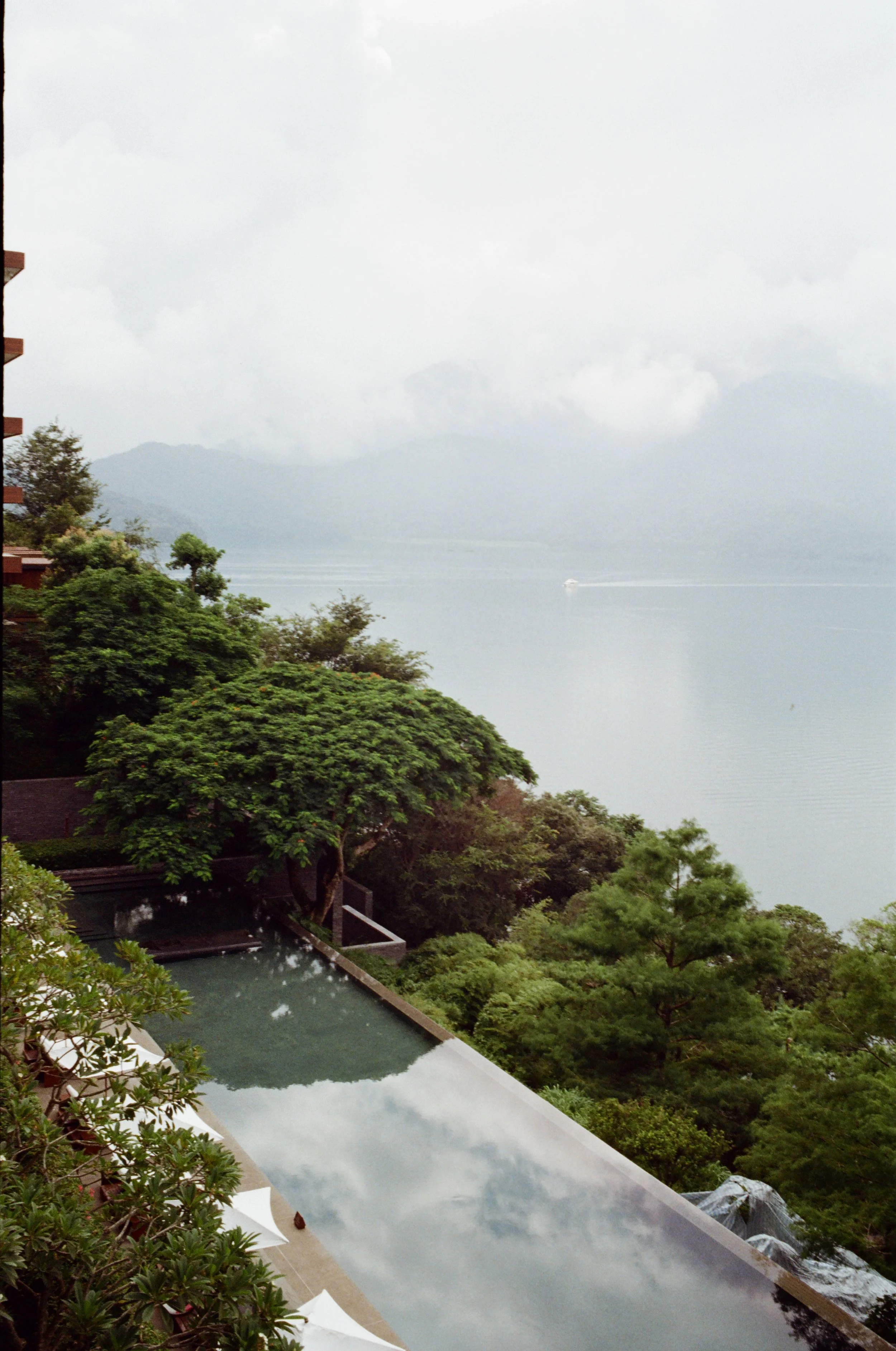 View from a balcony overlooking an infinity pool, lush green trees, and a lake or ocean in the distance with cloudy skies.