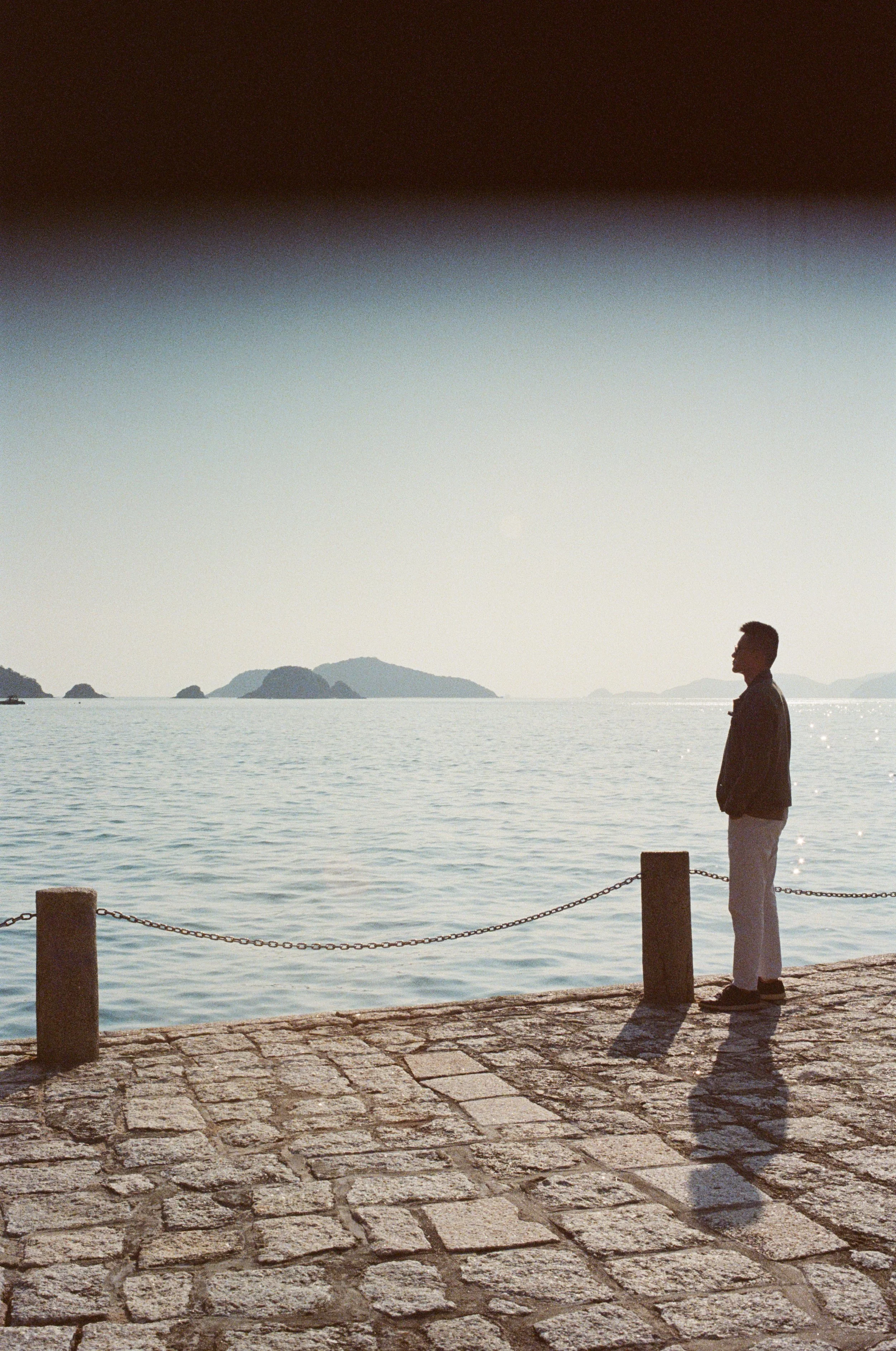 A person standing on a brick walkway by the water, looking out over the sea with islands in the distance, during sunset or sunrise.