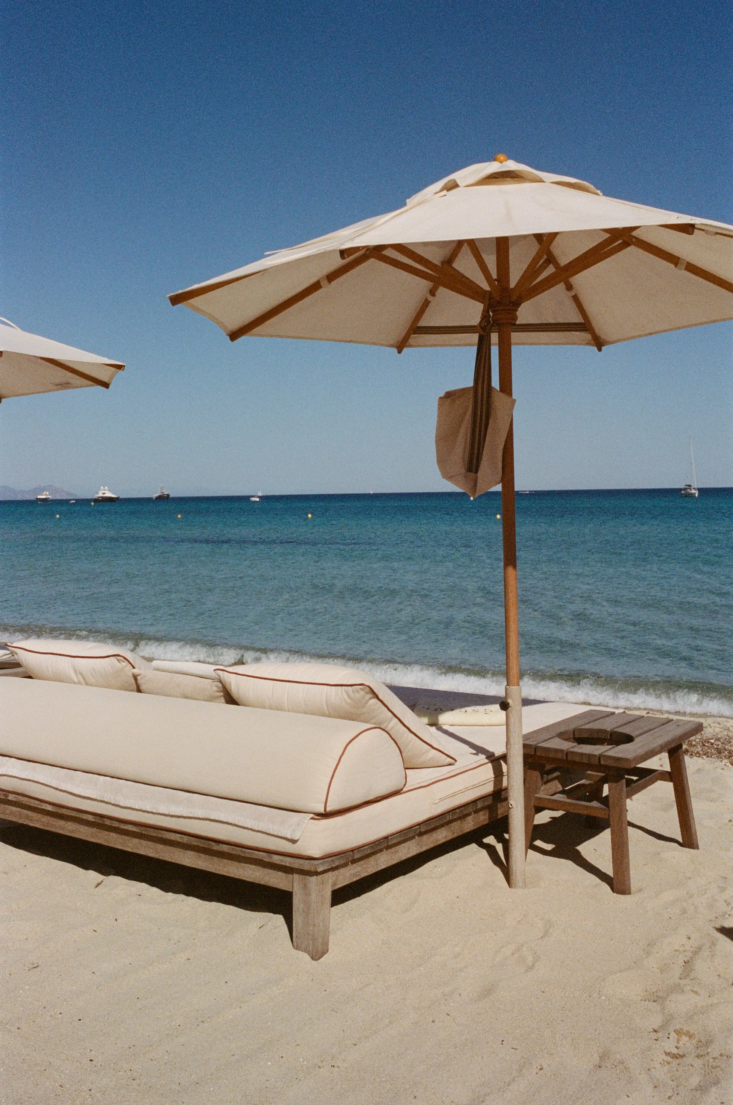 Beach scene with a lounge chair, side table, and large white umbrella on sandy shore overlooking the ocean with boats in the distance.