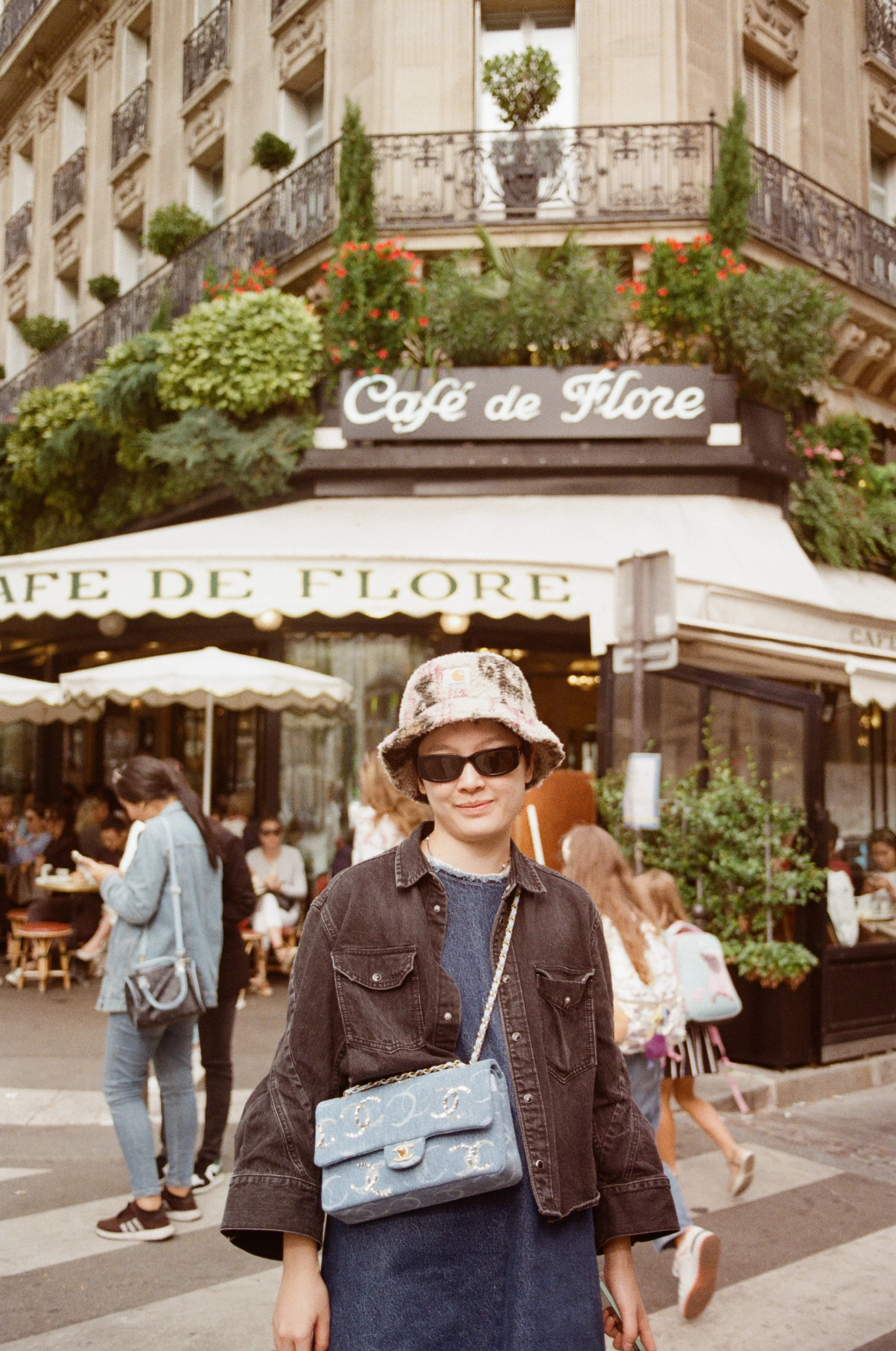 A woman wearing sunglasses, a bucket hat, a dark denim jacket, and a designer handbag stands in front of Cafe de Flore with outdoor seating and people walking by.