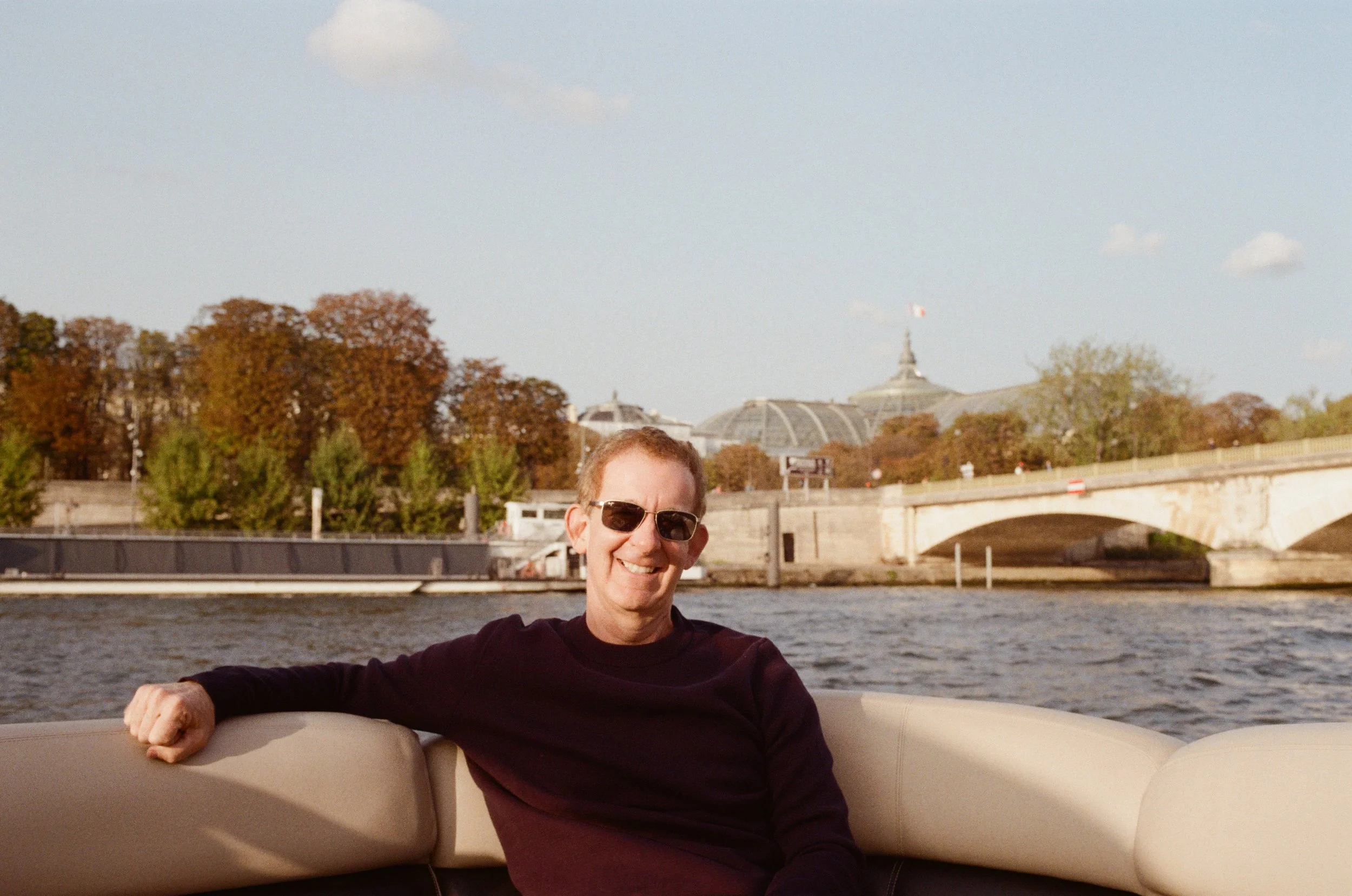 A smiling man wearing sunglasses and a dark sweater, sitting on a boat with a river and a bridge in the background, during daytime with trees and cloudy sky.