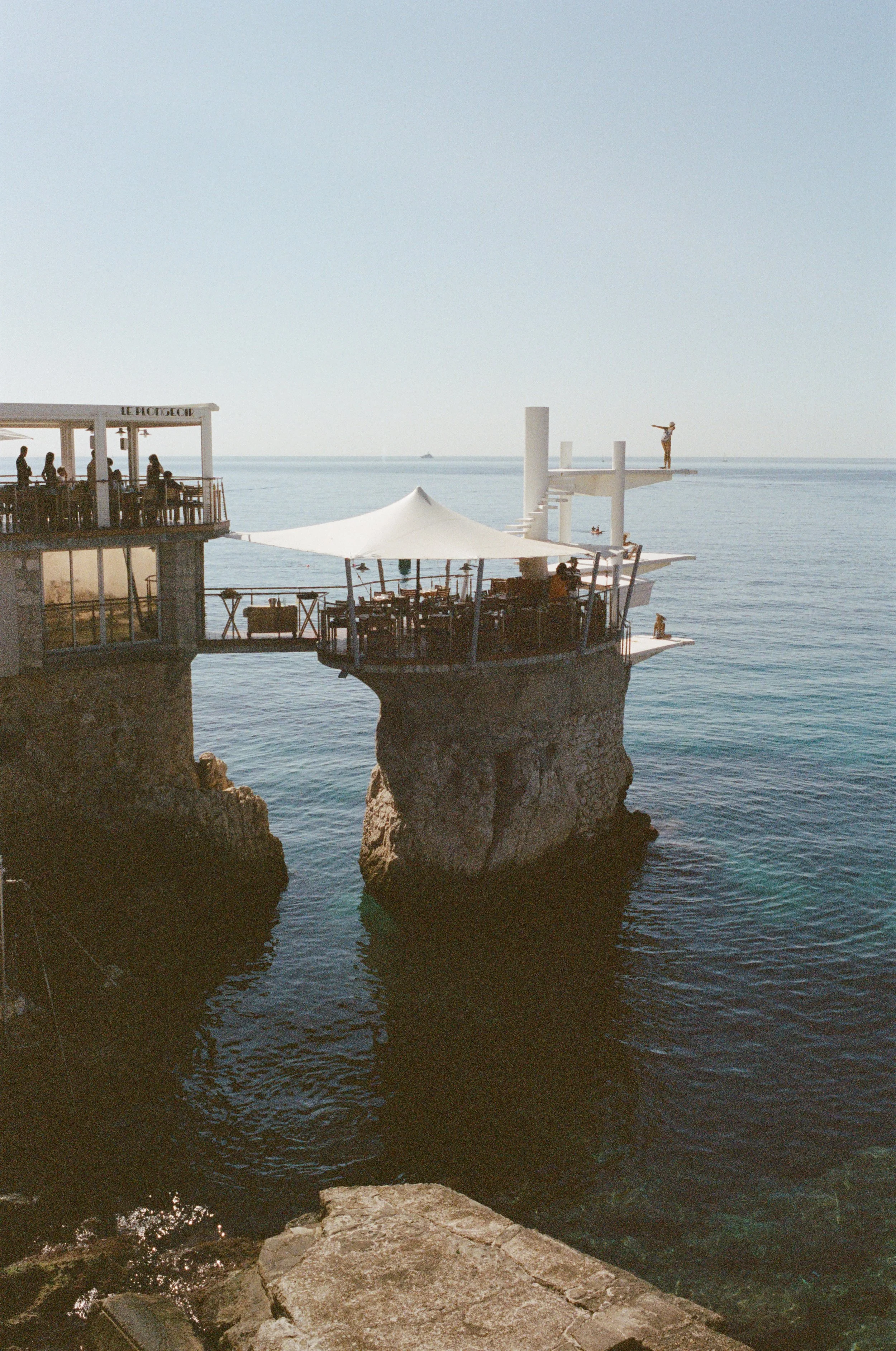 Cliffside restaurant with outdoor seating, a large white parasol, and a diving platform with a person preparing to jump into the sea, overlooking the calm ocean.