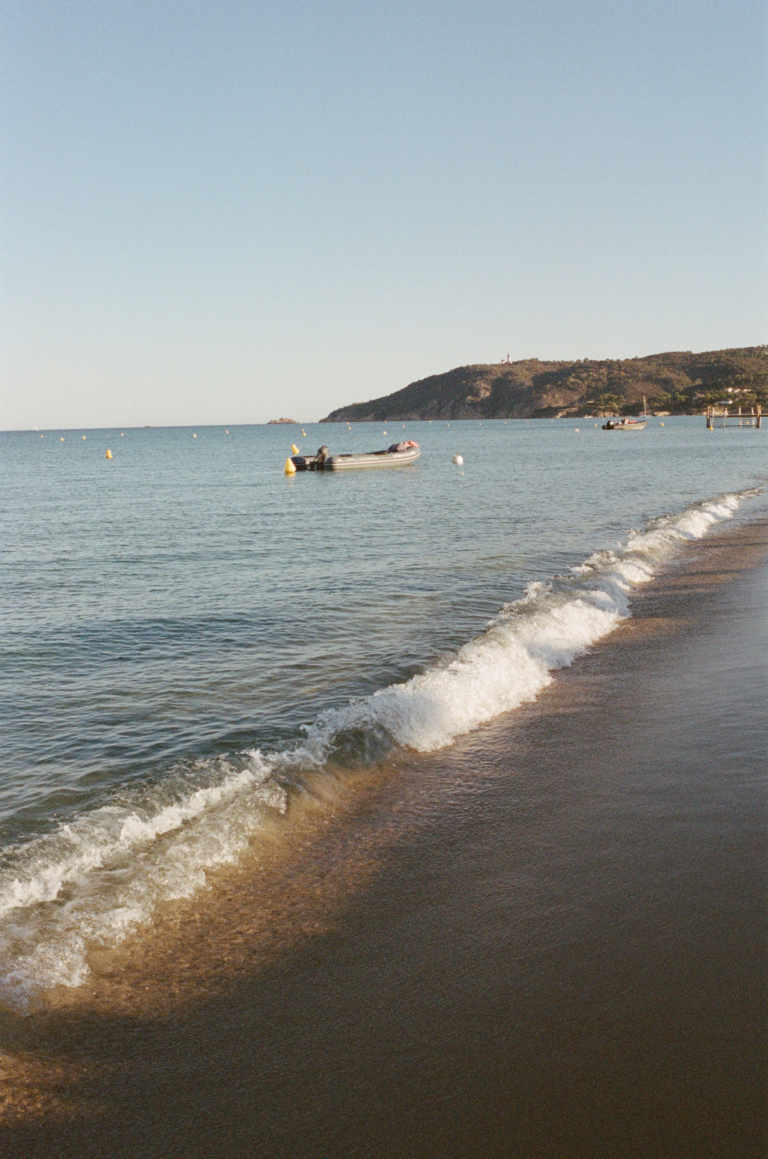 Calm beach with gentle waves, small boats floating on water, hilly landscape in the background, clear sky.