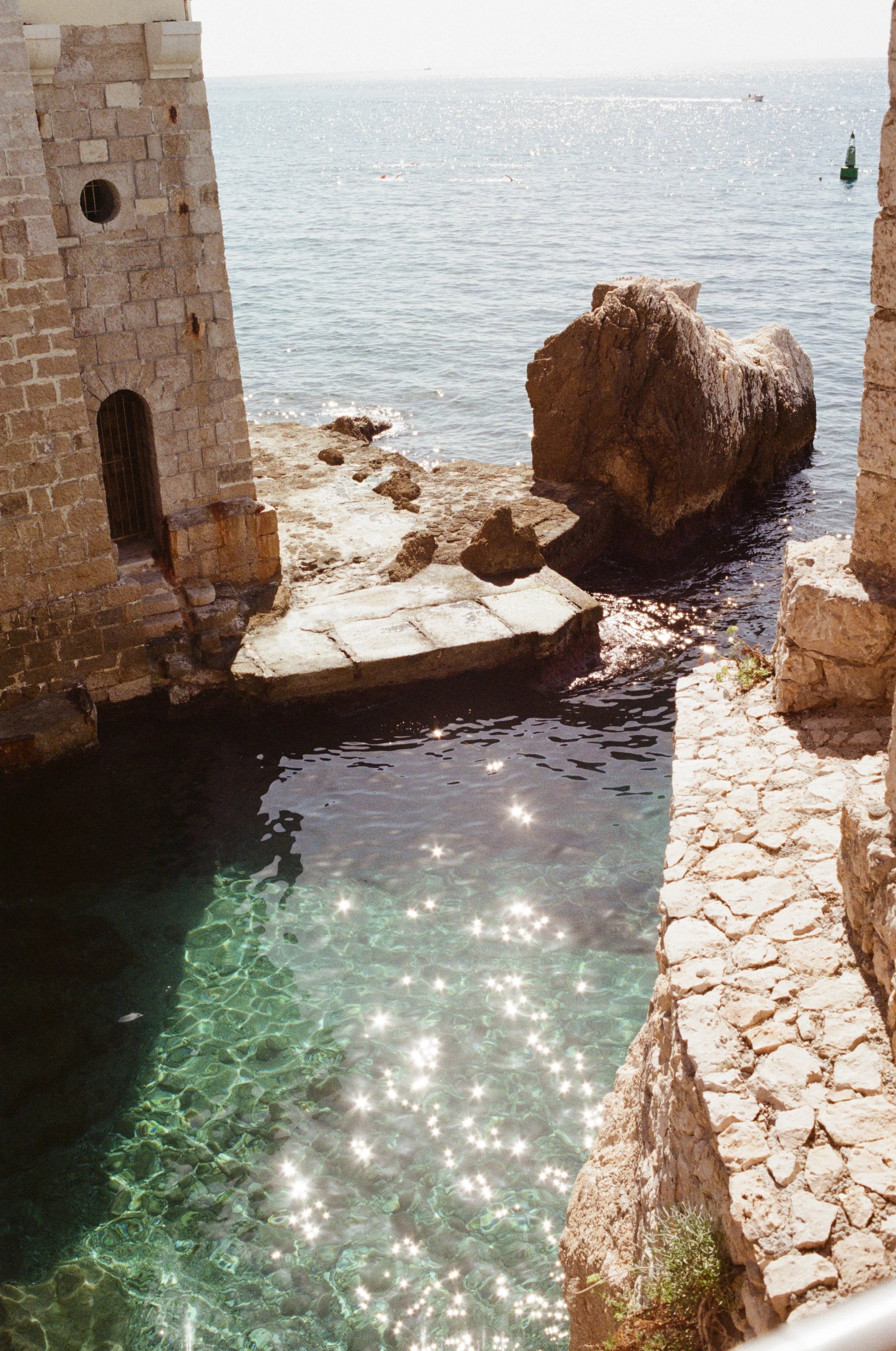 View of the sea through a stone archway with a small pool, large rocks, and a boat in the distance.