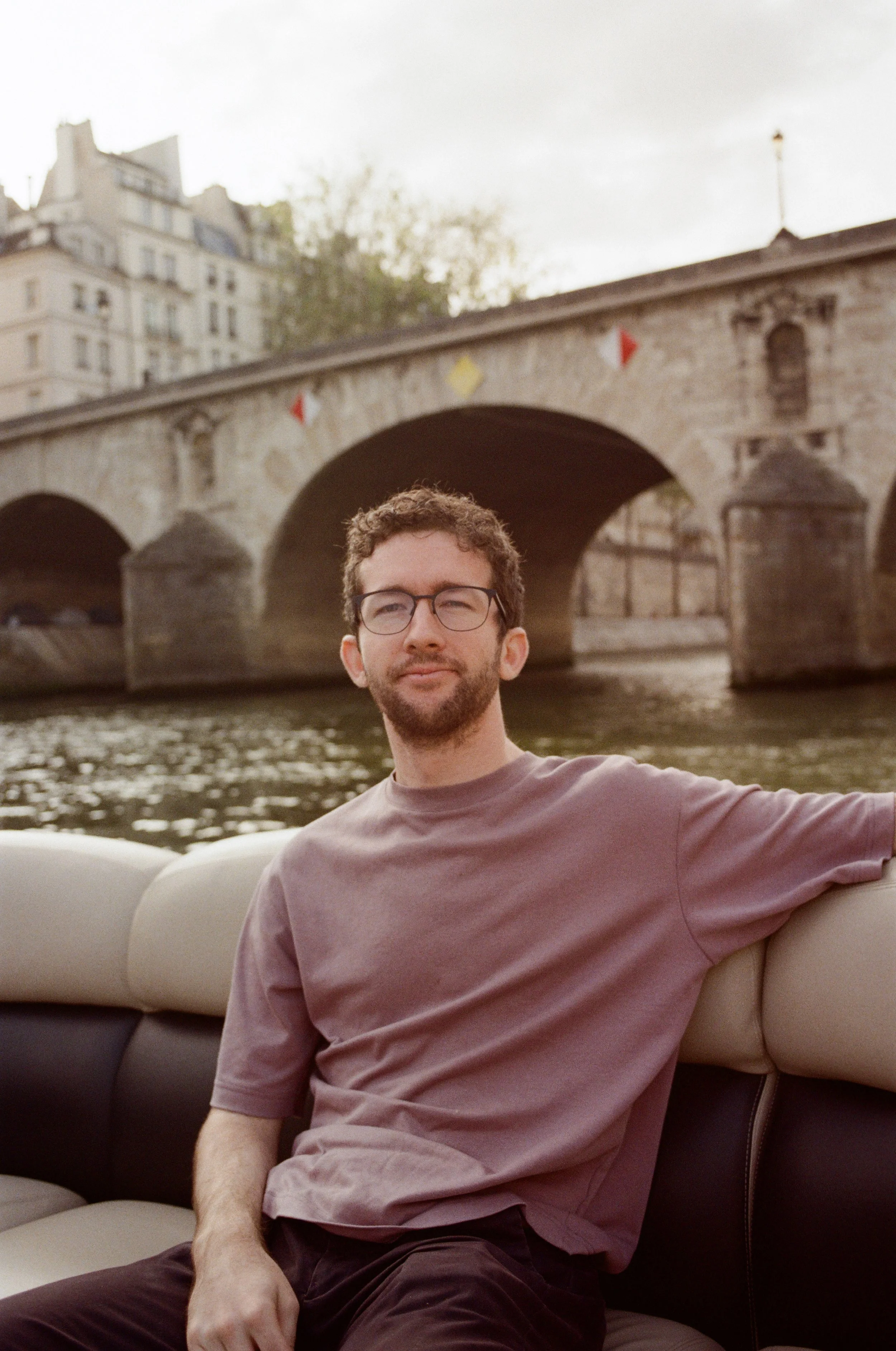 A man with glasses and a beard sitting on a boat near a river with an arched stone bridge in the background