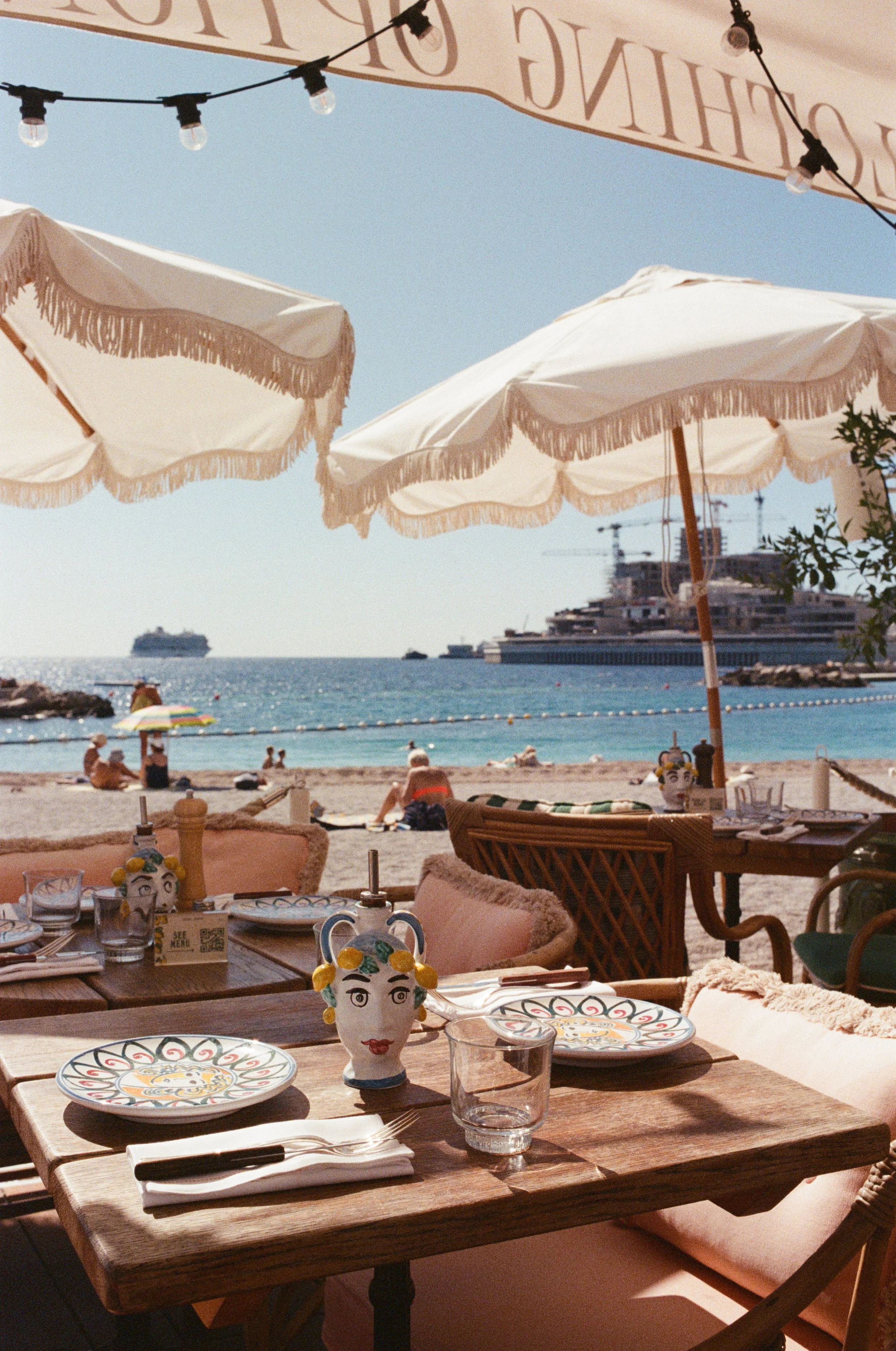 Beachside restaurant with tables and umbrellas, ocean and cruise ships in the background, sunny weather.
