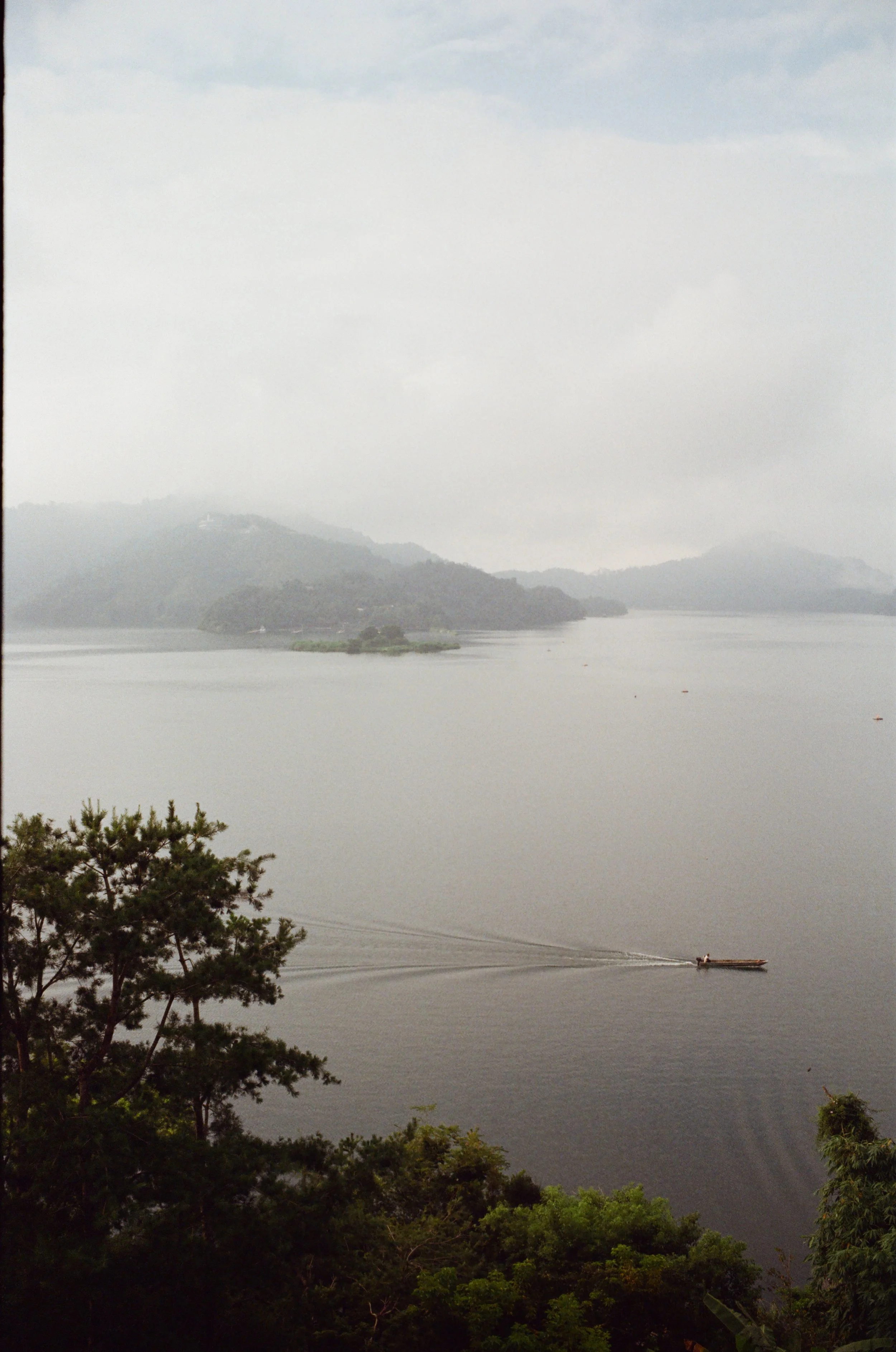 A river scene with a motorboat on calm water, lush green trees in the foreground, and misty hills in the background under a cloudy sky.
