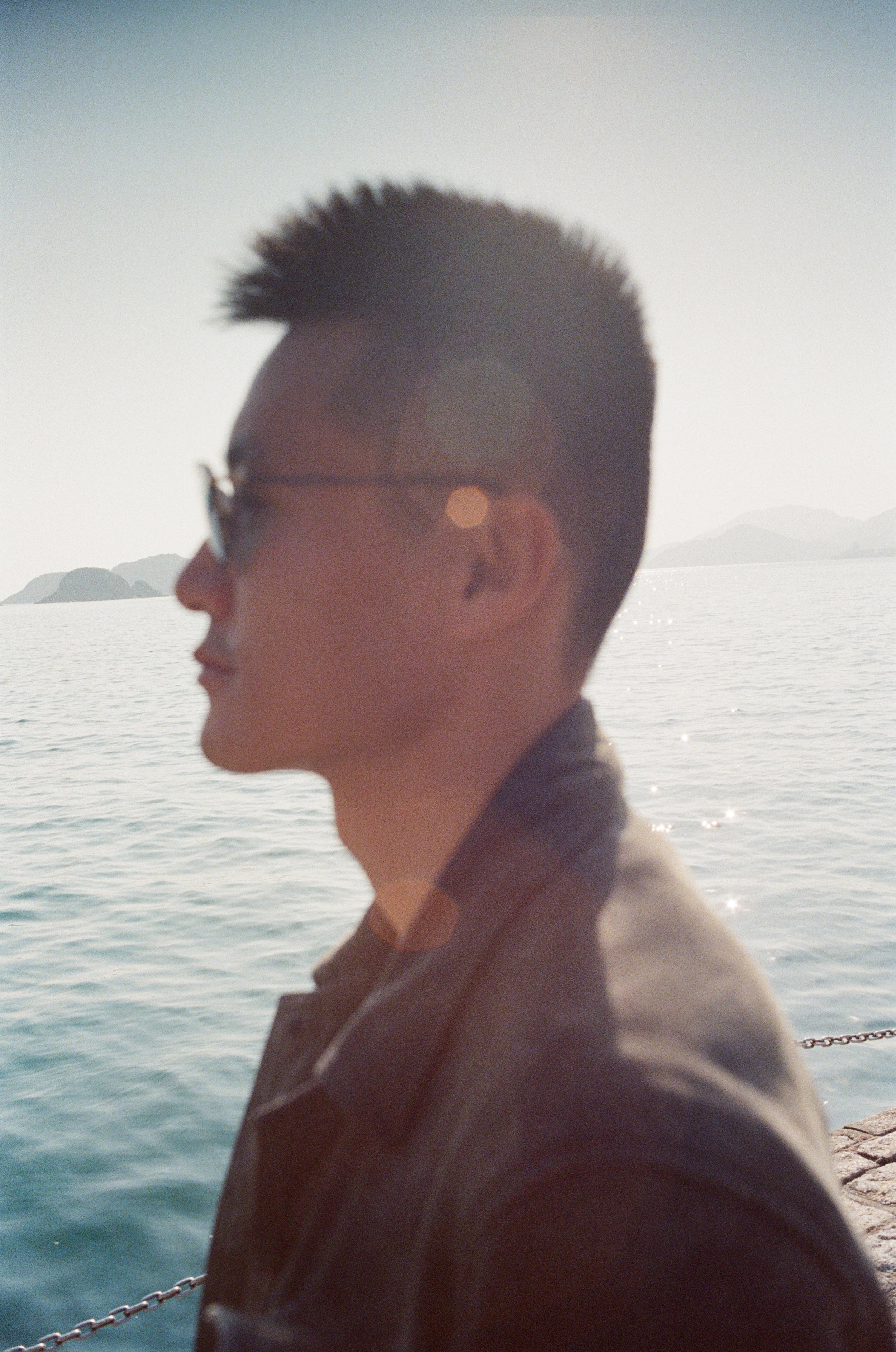 Profile of a young man wearing glasses, standing by a body of water with islands in the distance, during sunset.