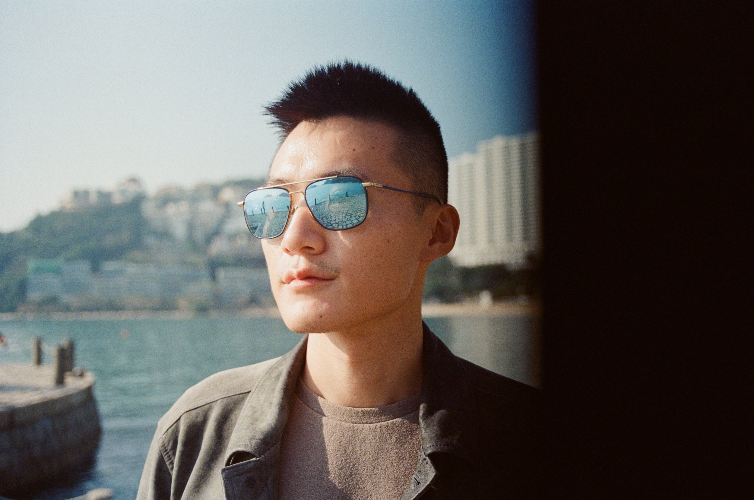 Young man with spiked hair and sunglasses looking to the side near a waterway with high-rise buildings in the background.