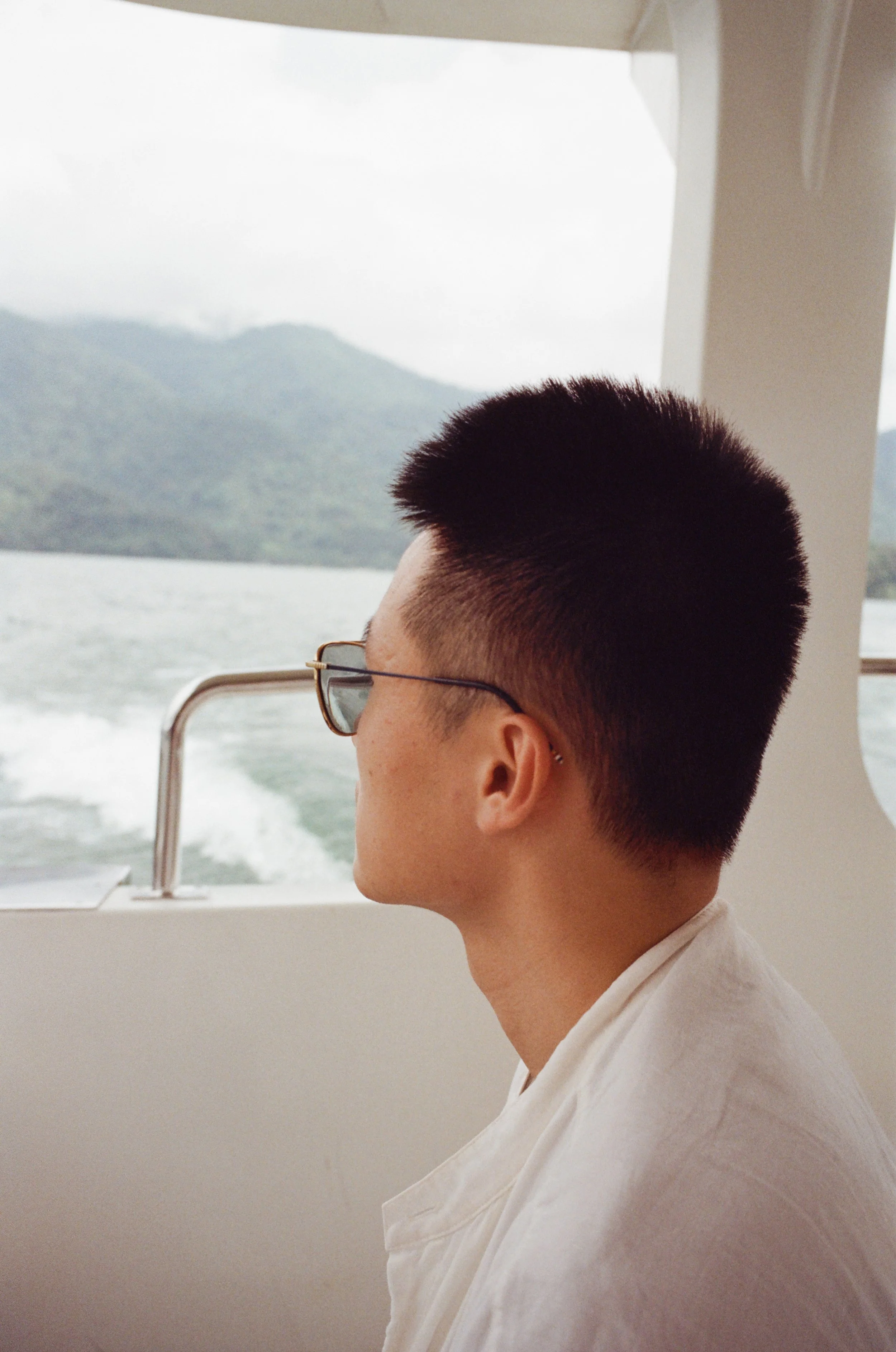 A person with short hair, glasses, and a white shirt looking out from the side of a boat, with a water and mountain landscape in the background.