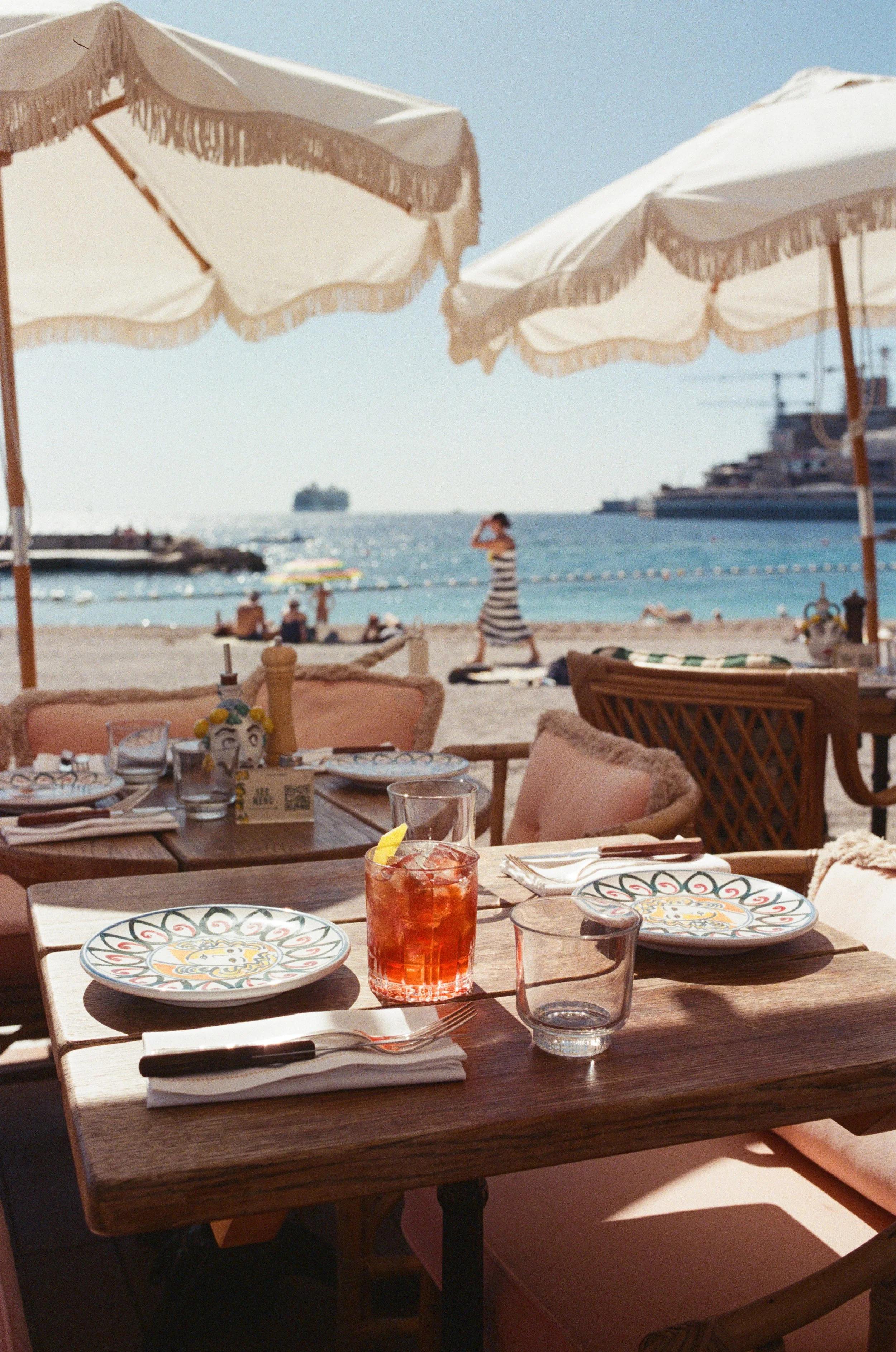 A seaside restaurant table set with plates, glasses, and cutlery, shaded by large white umbrellas, looking out onto a sandy beach with people relaxing and a person walking along the shore, and ships in the water.