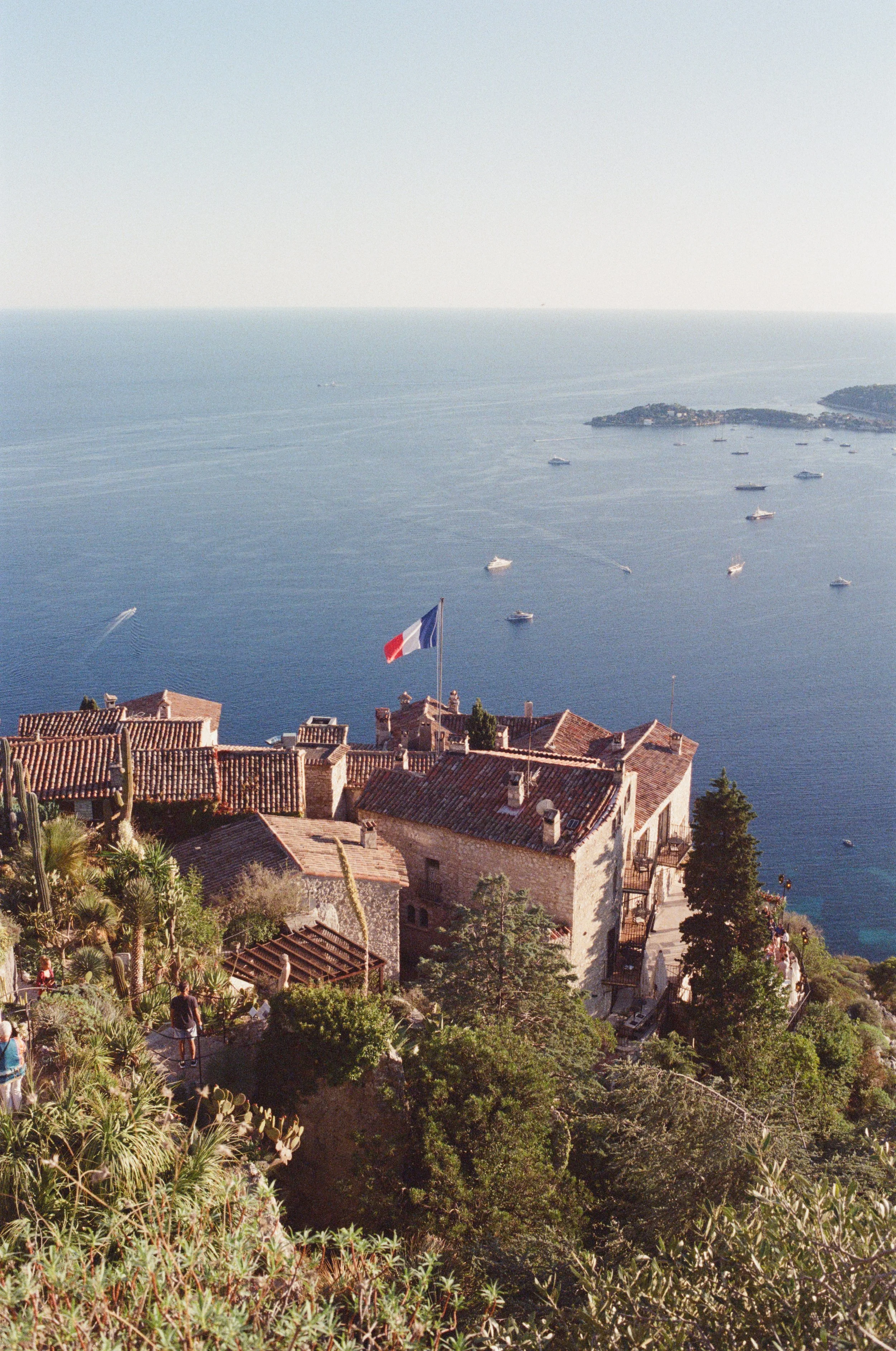 Coastal scene with buildings featuring red-tiled roofs and a French flag in the foreground, overlooking the sea with boats and a distant island.