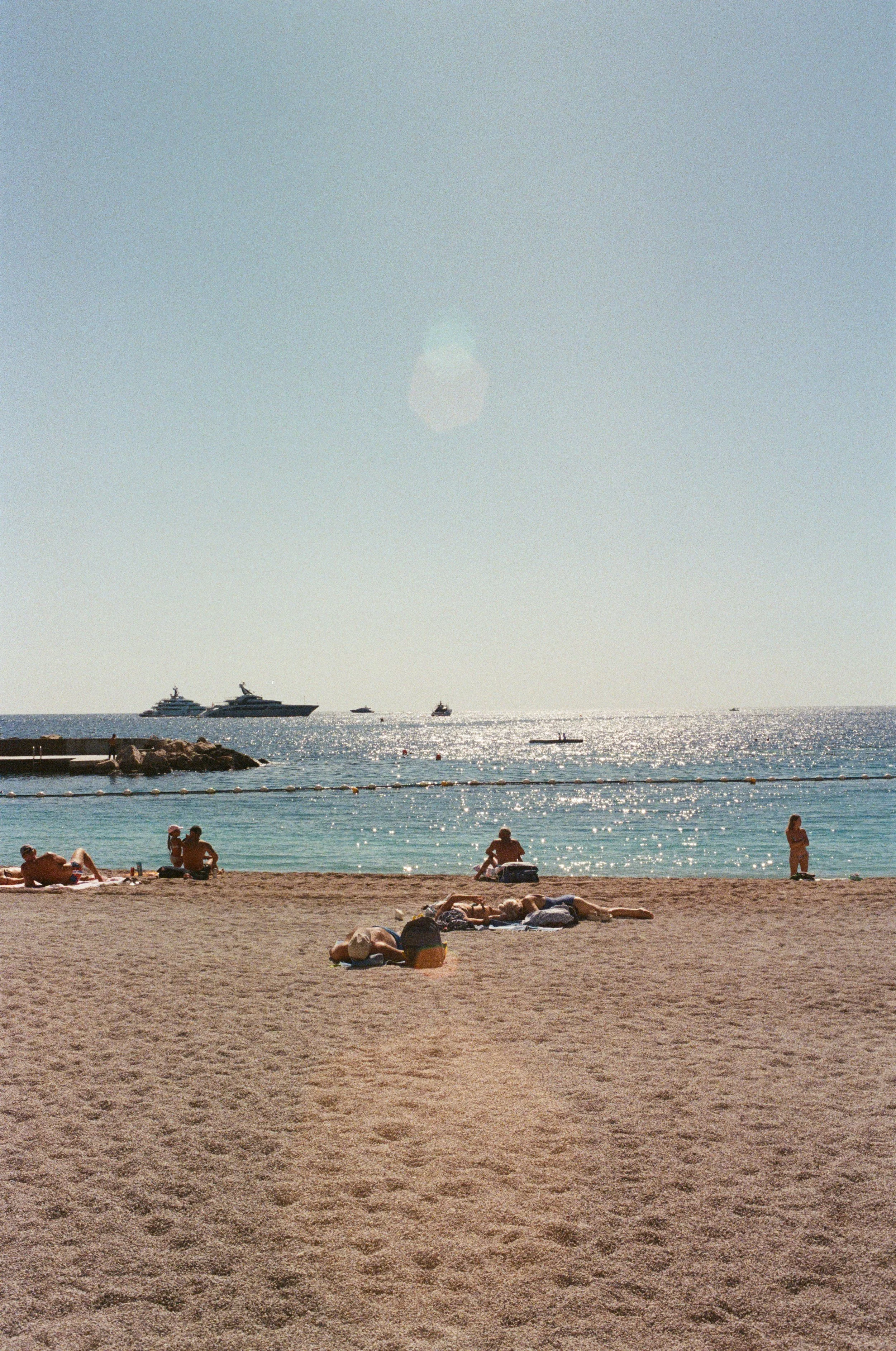 People relaxing on a sandy beach with boats and yachts in the water, and a clear sky in the background.