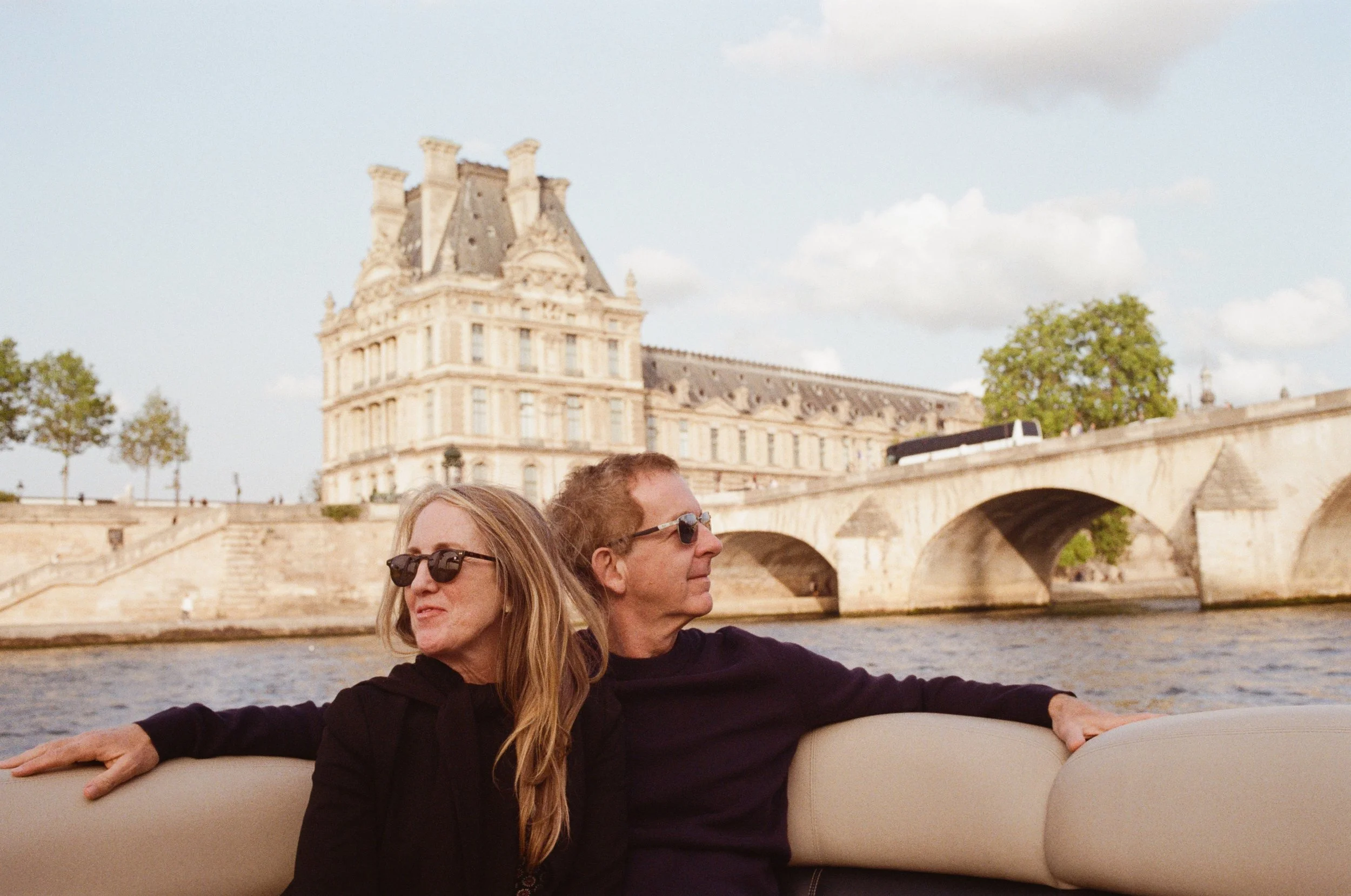 A woman and man wearing sunglasses are sitting on a boat with a view of a historic building and bridge over a river in Paris, France.