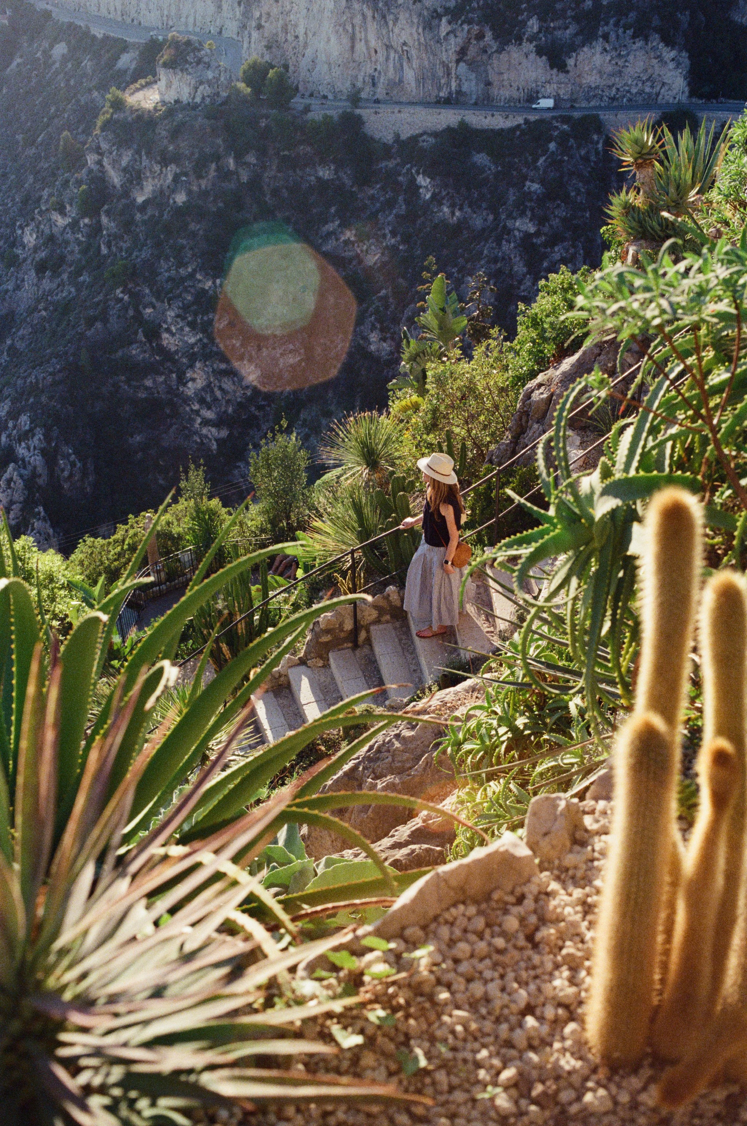 A woman wearing a straw hat, black top, and long skirt stands on stone steps surrounded by desert plants, overlooking a canyon with a winding road and a vehicle in the distance.