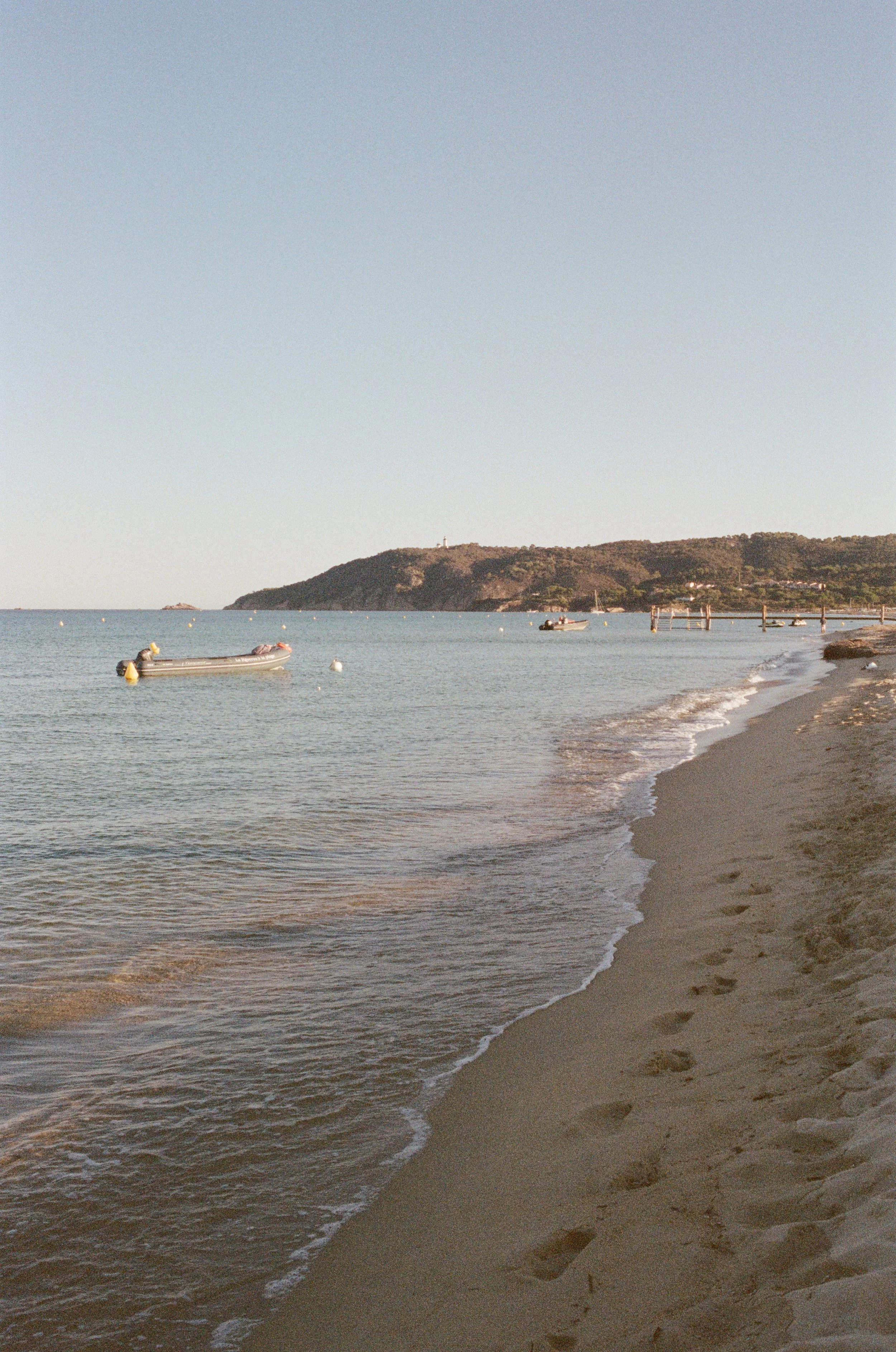A peaceful beach scene with calm water, a sandy shoreline with footprints, several moored boats, and a distant hillside under a clear sky.