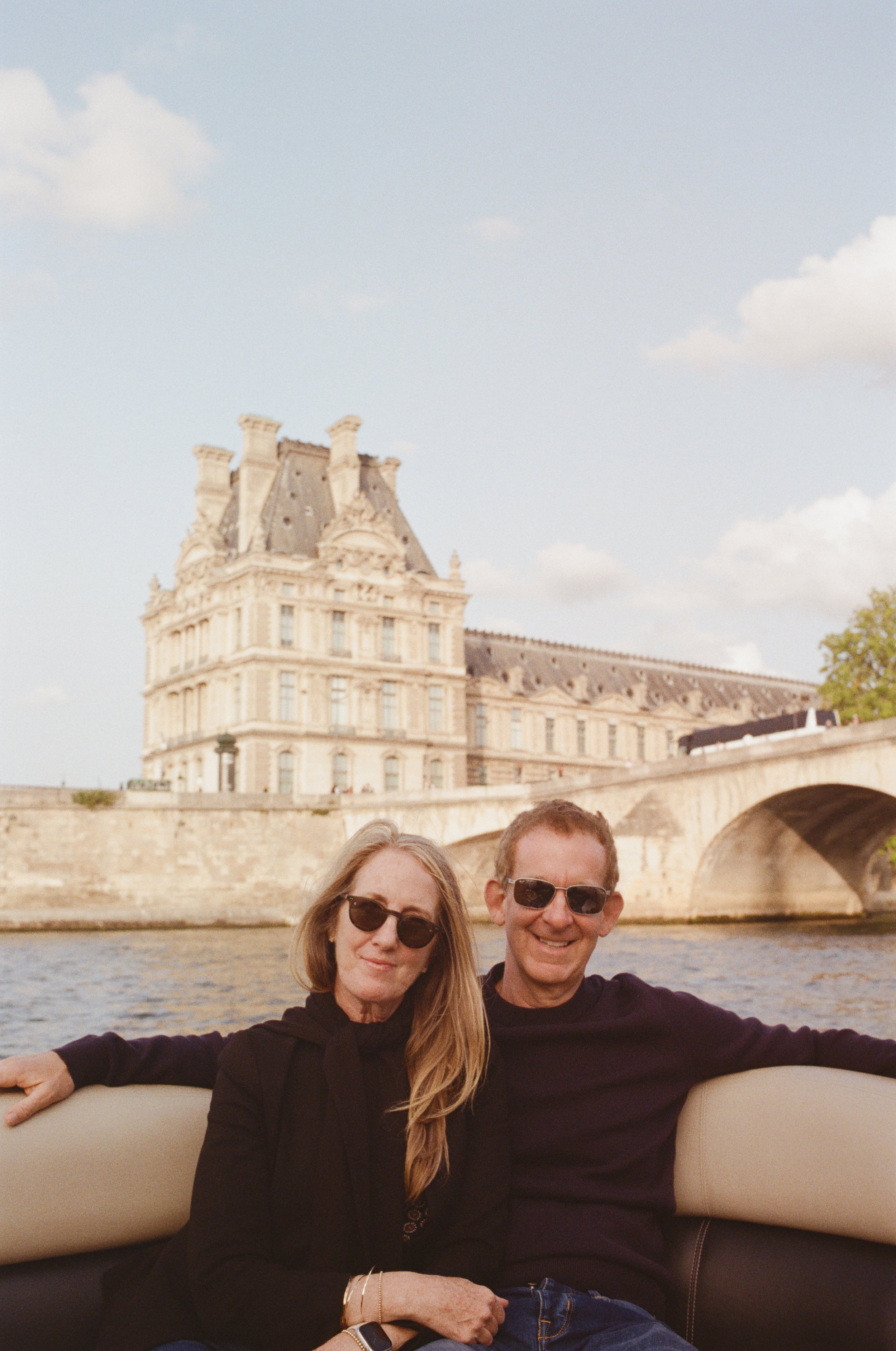 A middle-aged couple wearing sunglasses sitting on a boat in front of a river with a historic building and bridge in Paris, France, visible in the background.