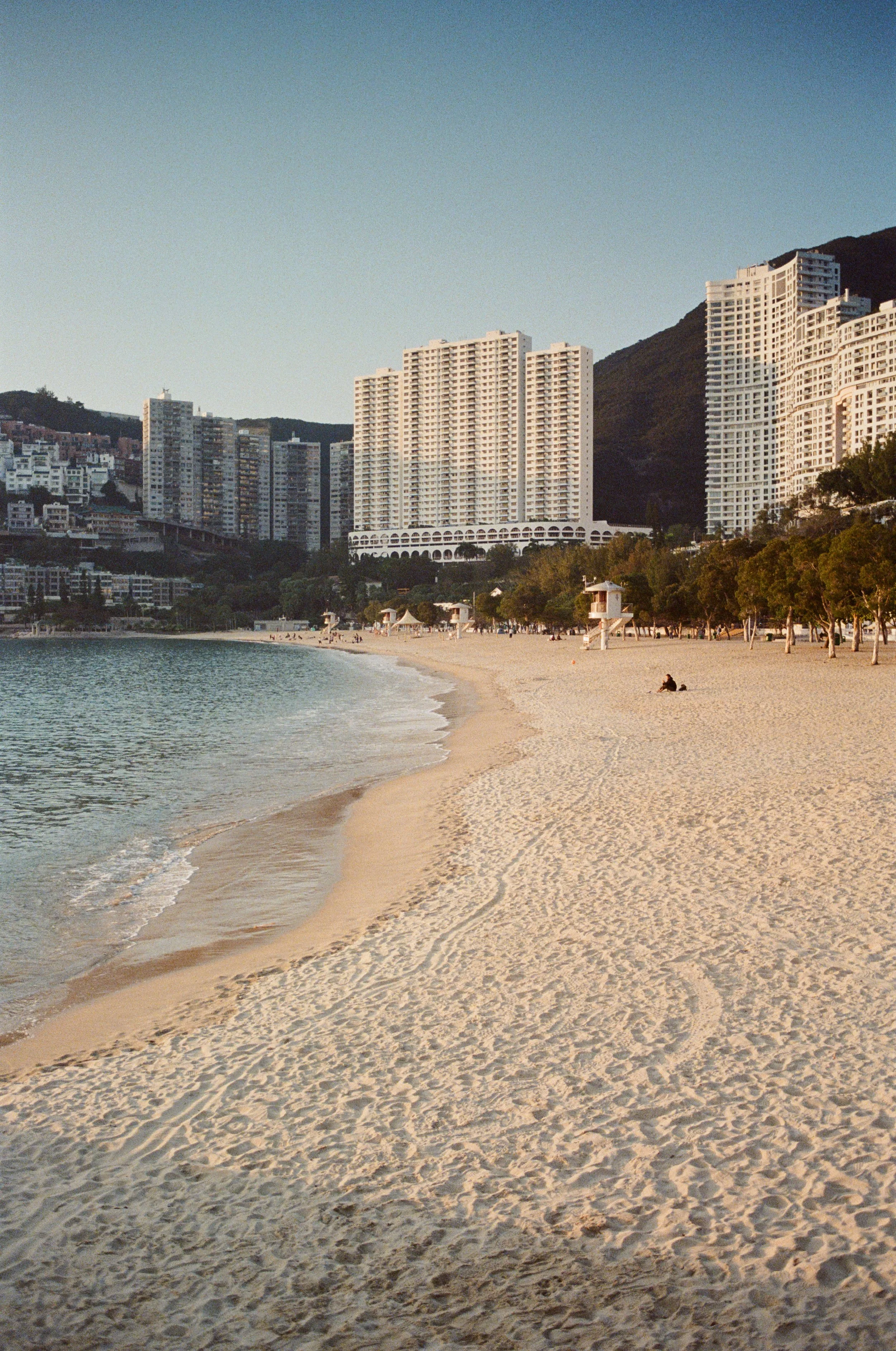 A beach with white sand, calm water, and a cityscape of high-rise buildings in the background. There are a few people on the beach and some trees along the shoreline.