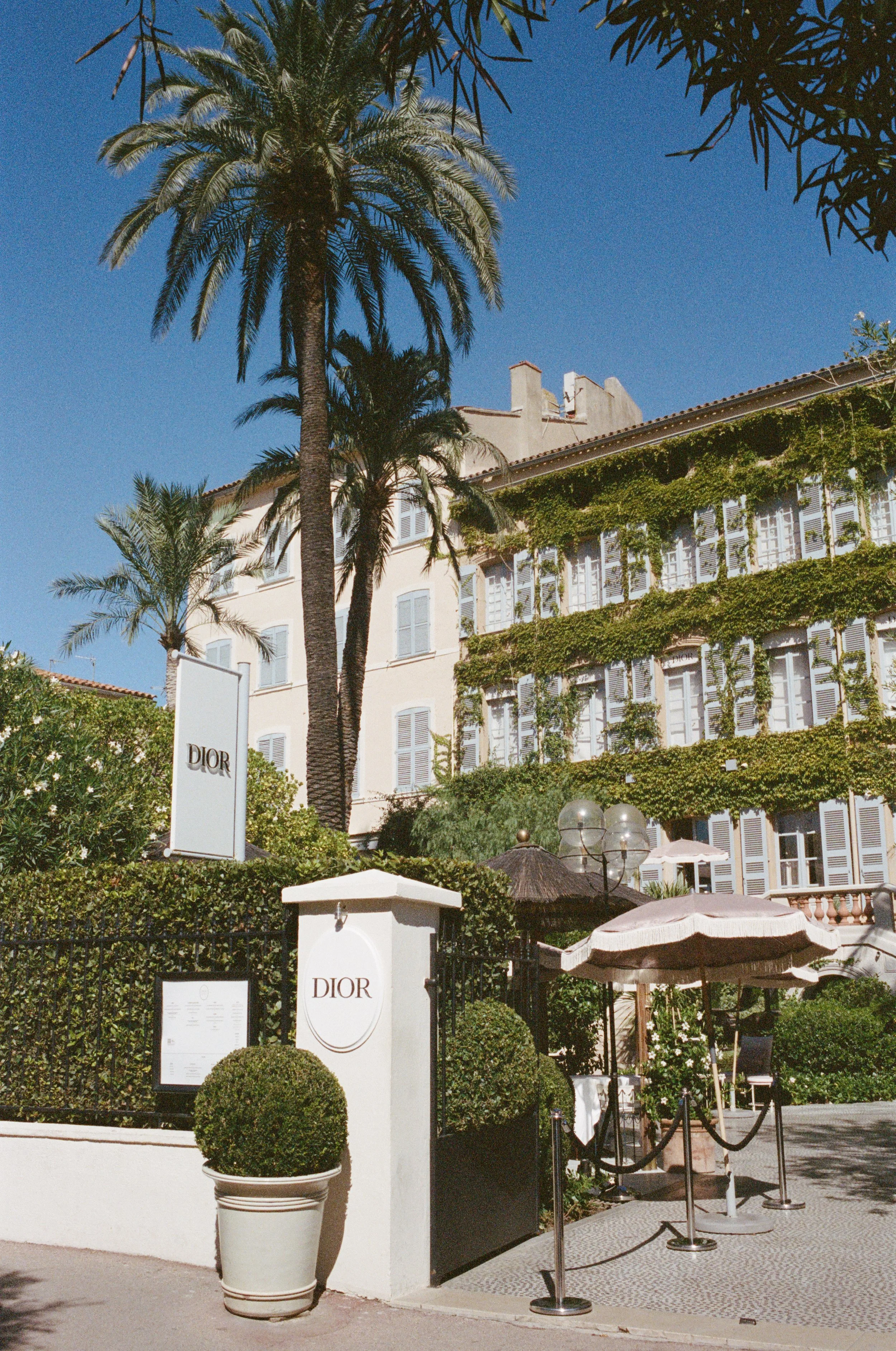 Exterior view of the Dior boutique in a sunny location, featuring a white building covered in green vines, palm trees, and outdoor seating with umbrellas.