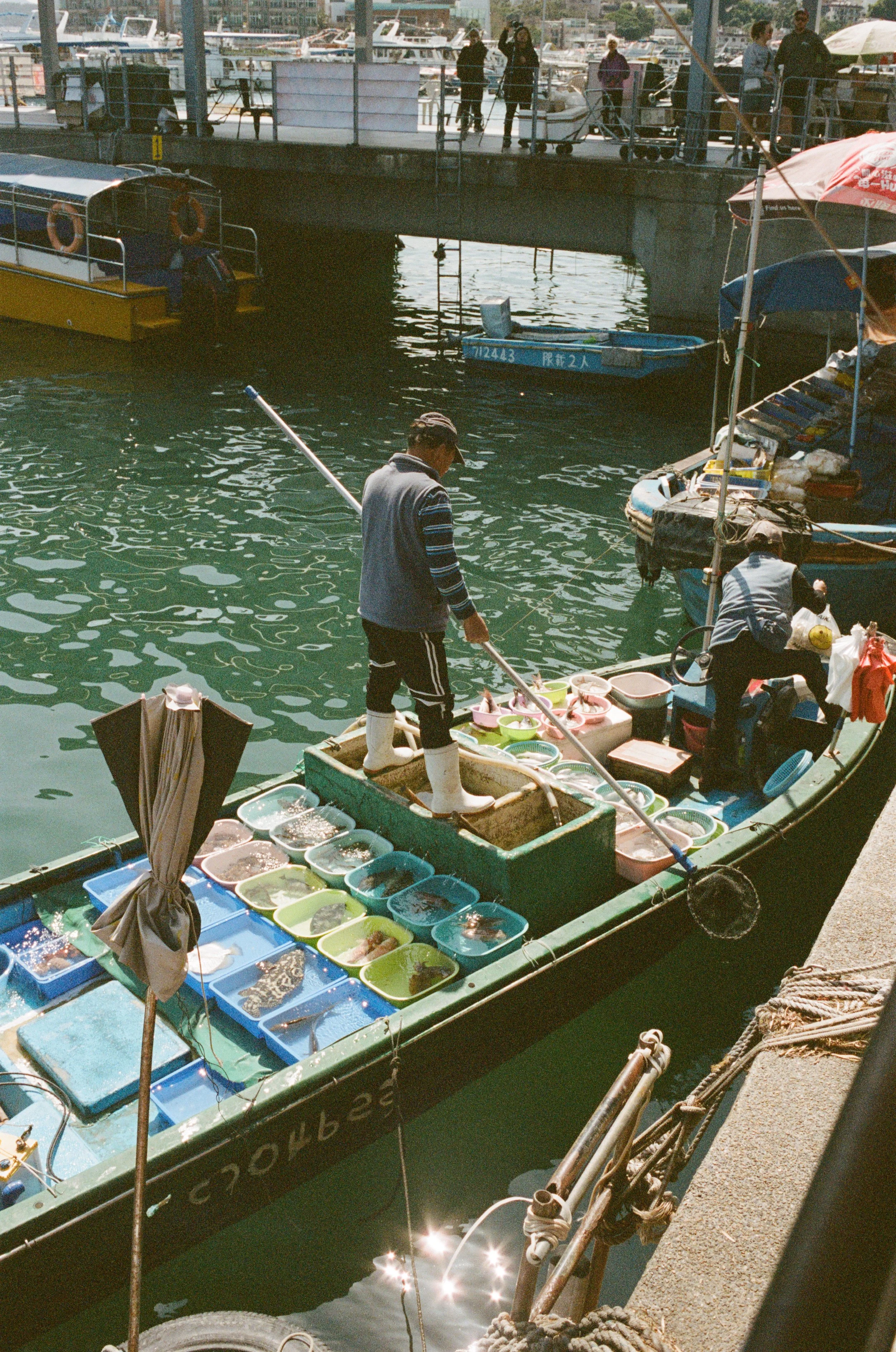 A boat at a marina with two men selling seafood, including fish and other ocean creatures, displayed in plastic containers.