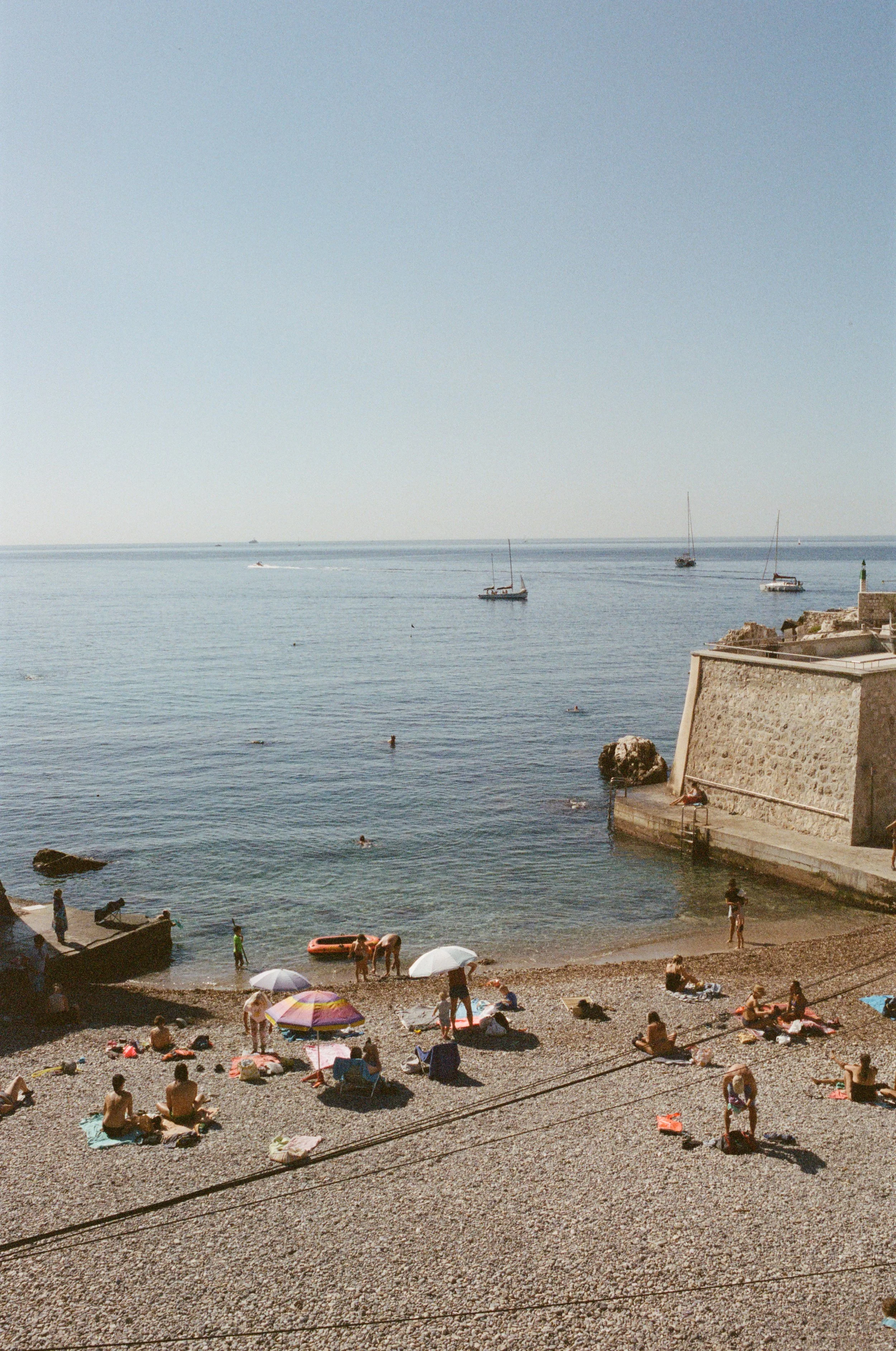 People relaxing and swimming at a pebble beach near the sea, with sailboats on the water and clear sky above.