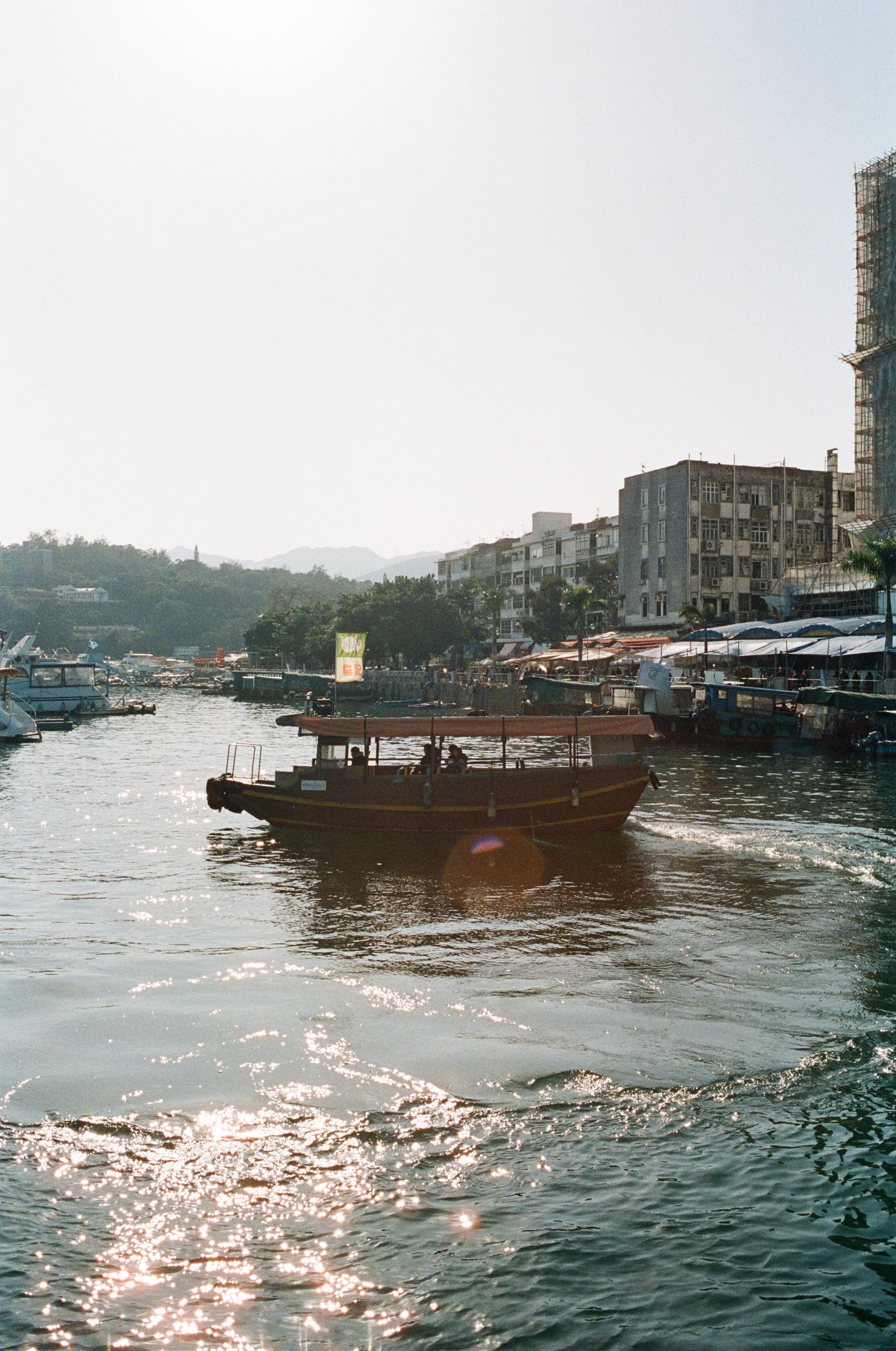 A boat sailing on a waterfront, with buildings and boats in the background, and sunlight reflecting on the water.