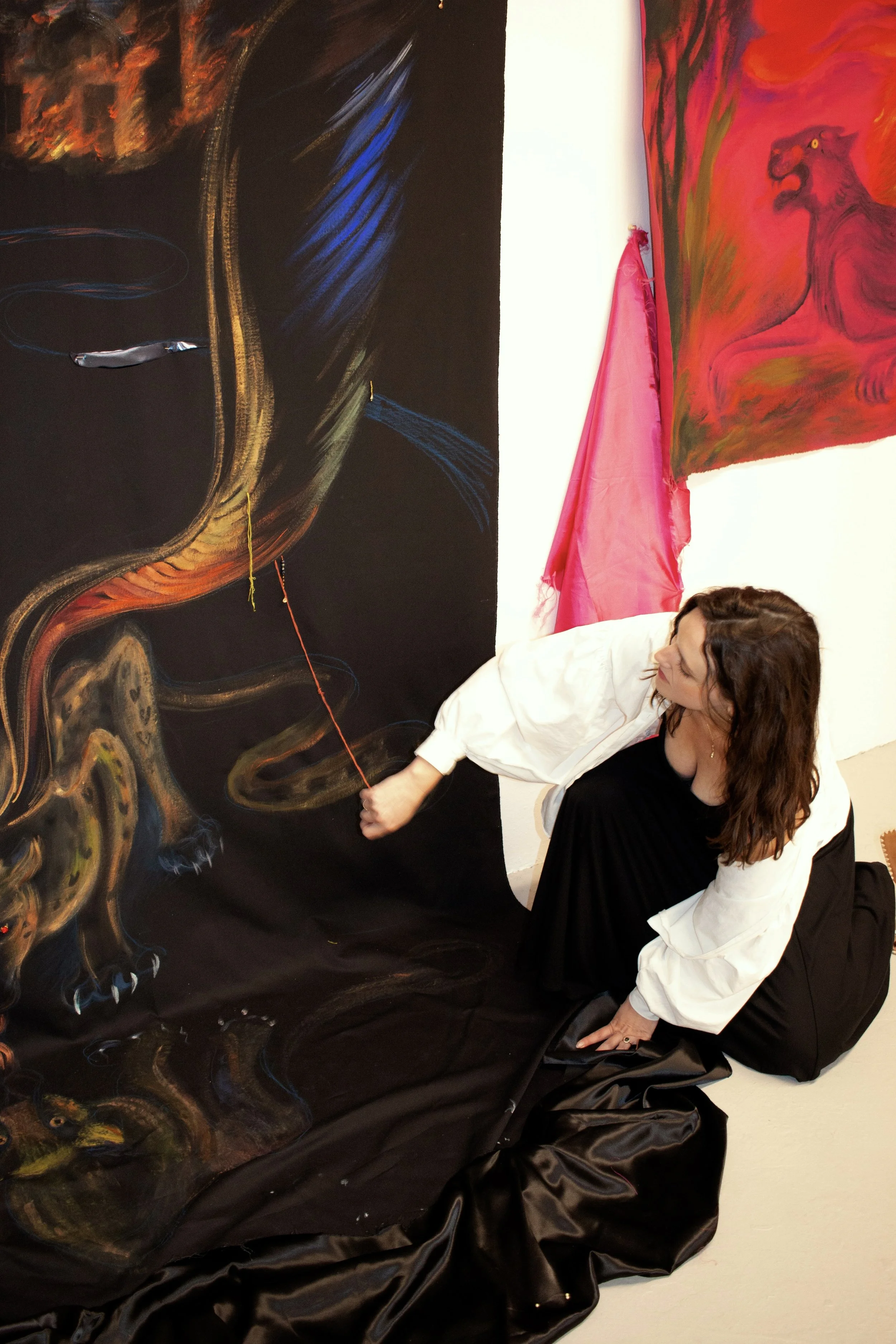 A woman with brown hair wearing a white jacket and black dress kneels on the floor, drawing or painting on a large black canvas that features a colorful abstract dragon. The background has white walls with colorful artwork, including a pink fabric decoration and a red painting with a creature that resembles a lion or a dog.