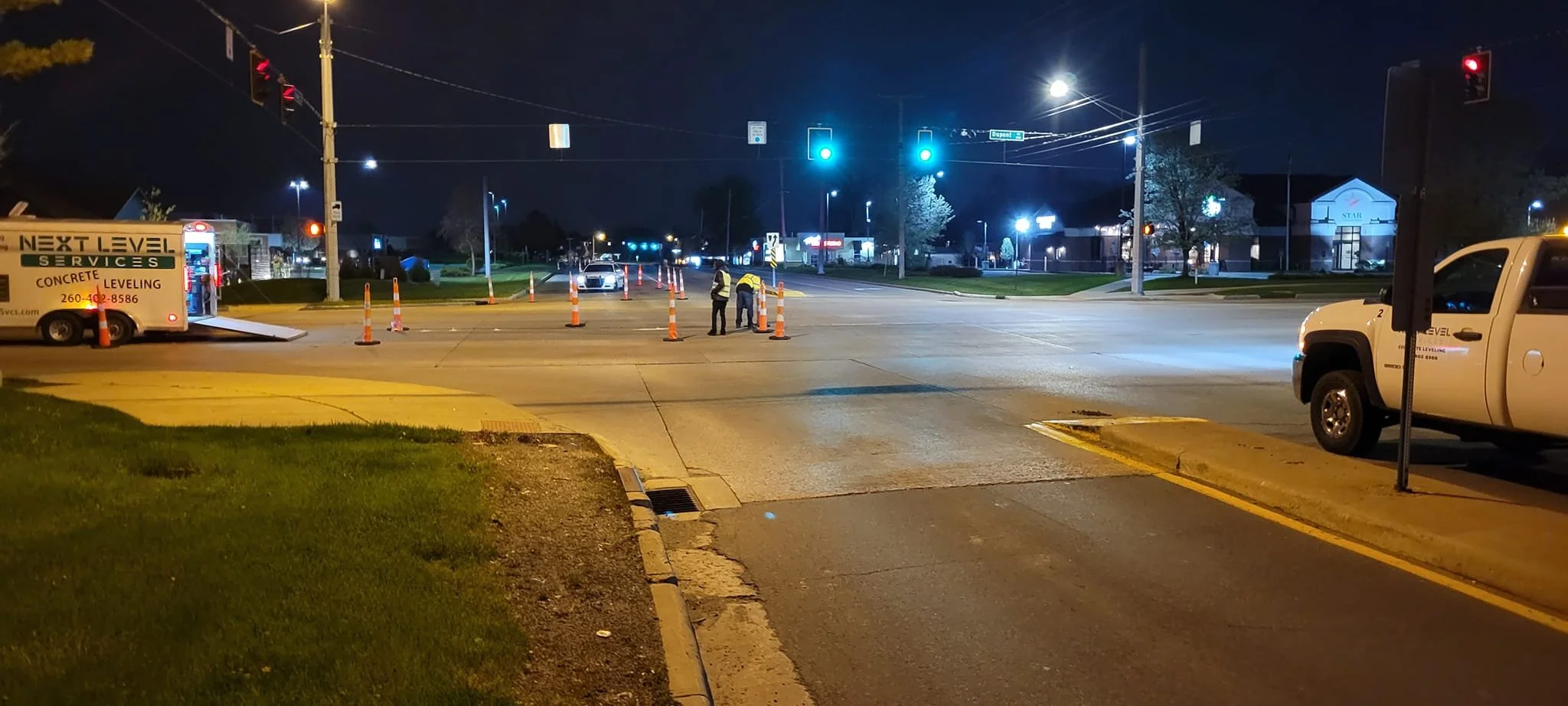 Nighttime traffic intersection with workers setting up traffic cones, a service van on the left, and a pickup truck on the right.