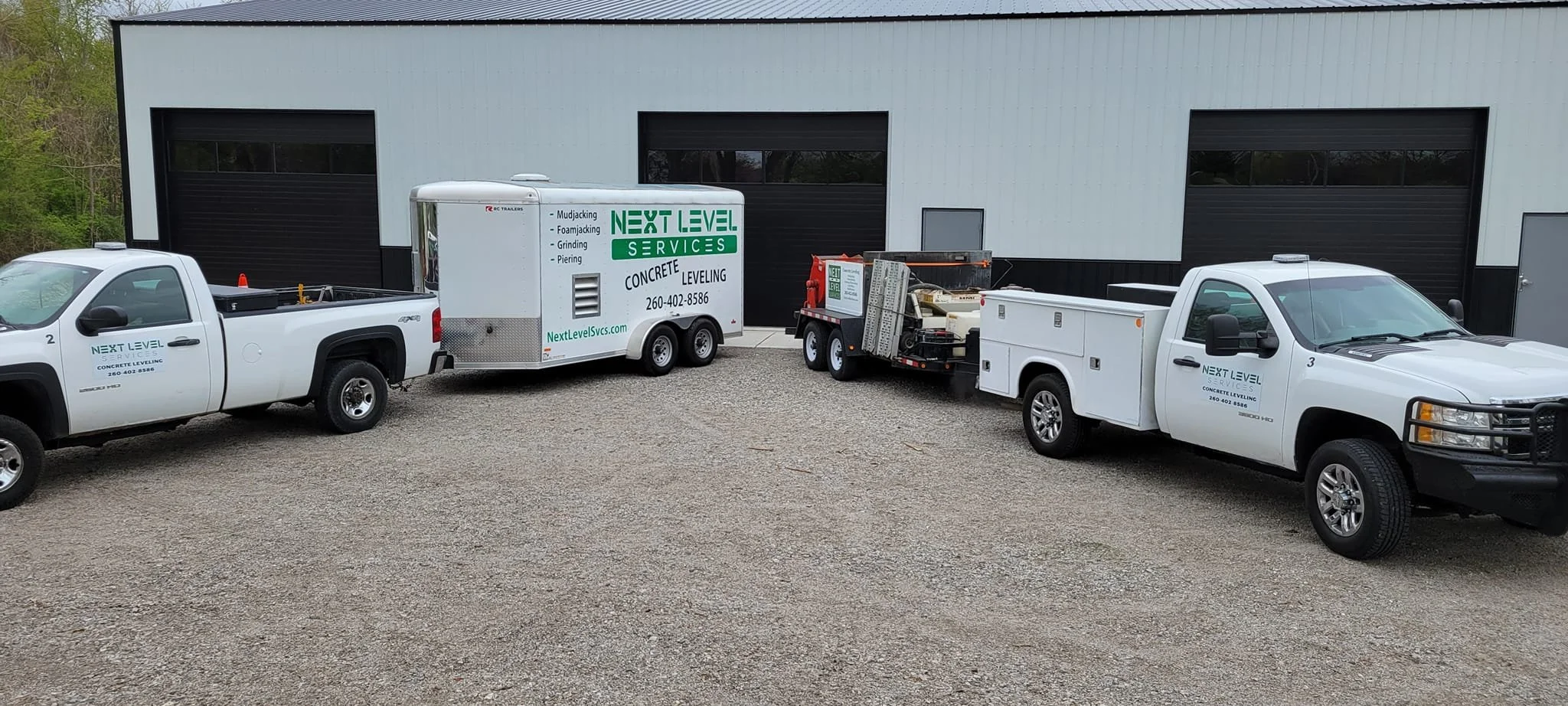 Two white service trucks and a trailer parked outside a industrial building with black and gray exterior walls and large black garage doors.