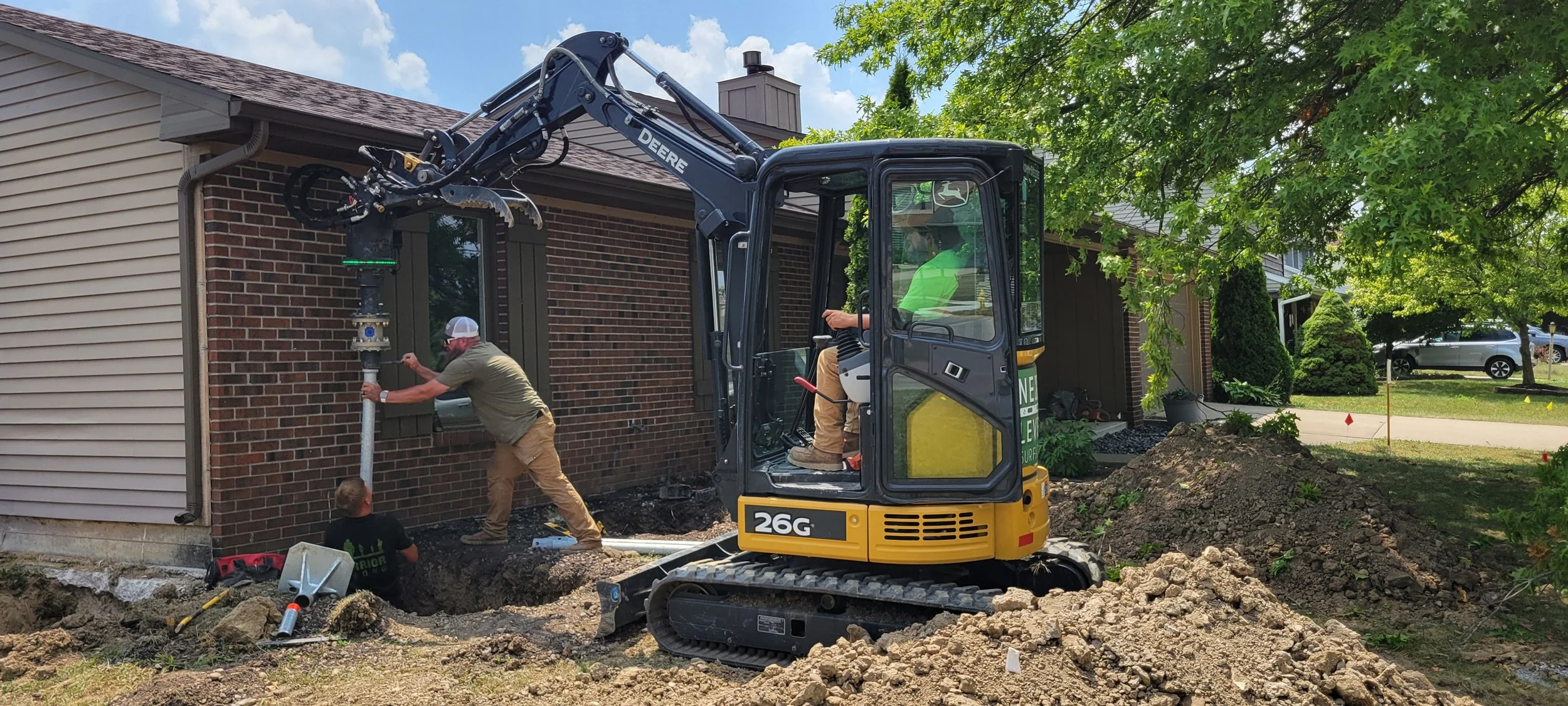 Construction workers installing a pipe near a brick house, using a small excavator on dirt and gravel in a residential yard with trees and a parked car in the background.