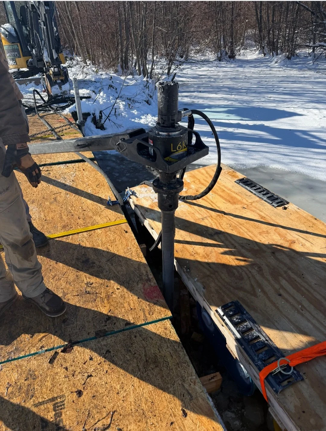 Construction site with a wood platform on ice and snow, a pile driver, and a person in gloves and work clothes operating machinery outdoors in winter.