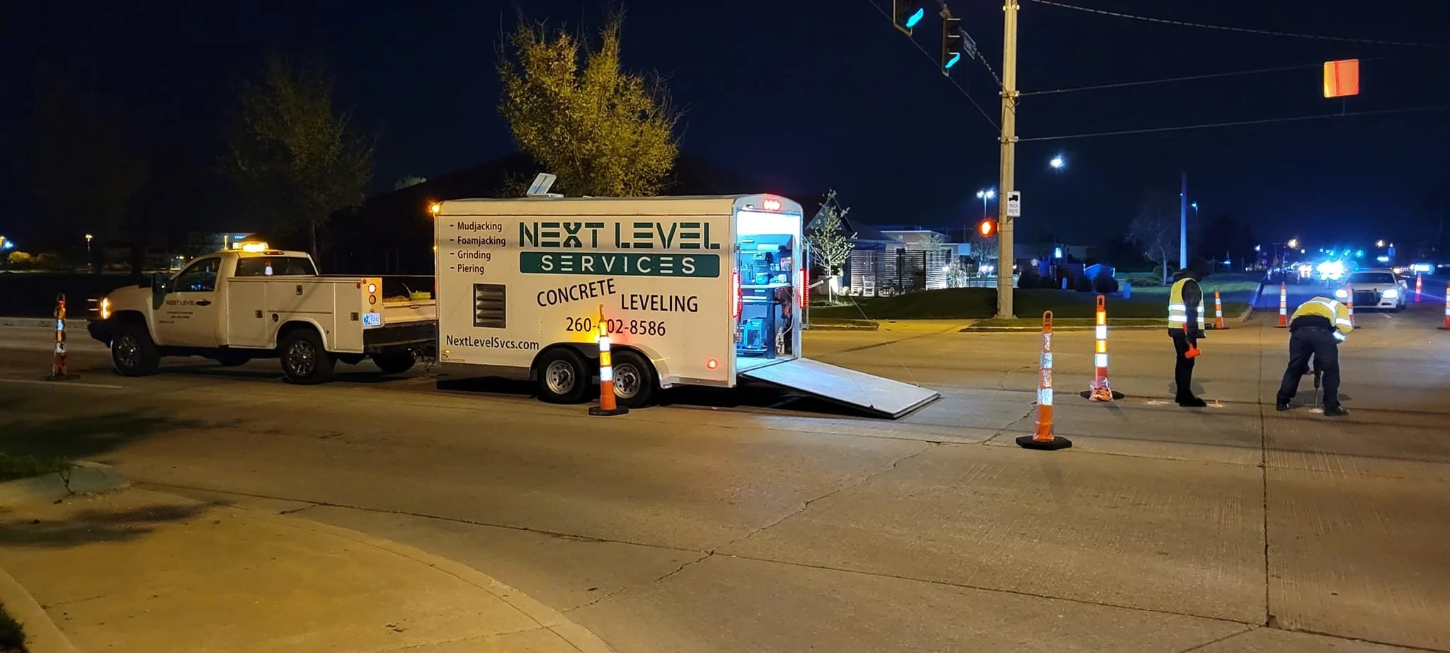Nighttime street scene with a construction or utility service truck labeled 'Next Level Services' and 'Concrete Leveling.' Two workers in reflective vests are working on the street, surrounded by orange traffic cones. In the background, police cars w