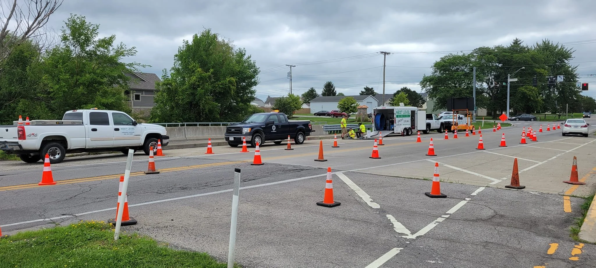 Road construction workers and traffic cones on a street. Utility truck and repair vehicles present. Overcast sky.