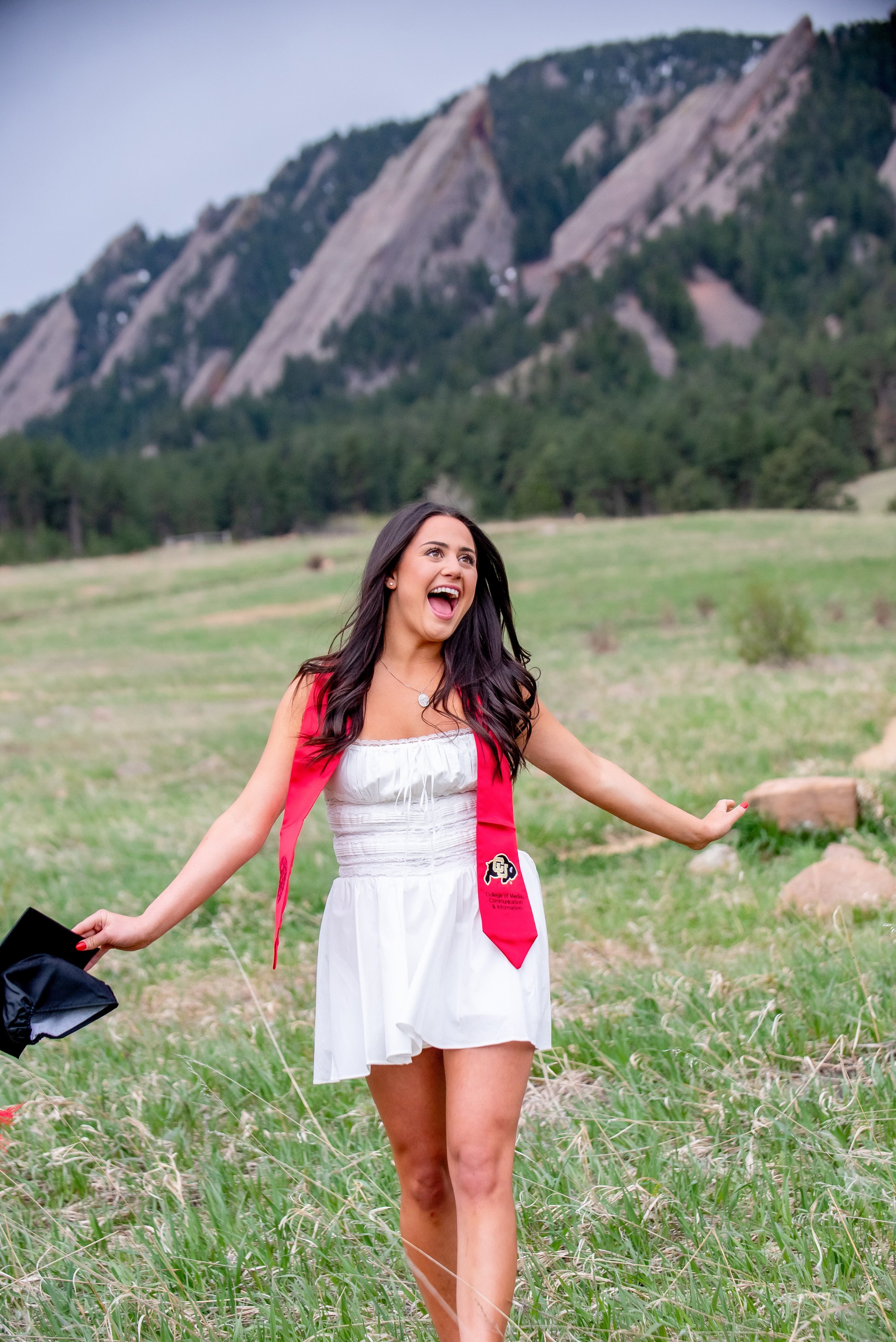 A young woman celebrating outdoors in a field, wearing a white dress and a red graduation stole, holding a graduation cap, with mountains and trees in the background.