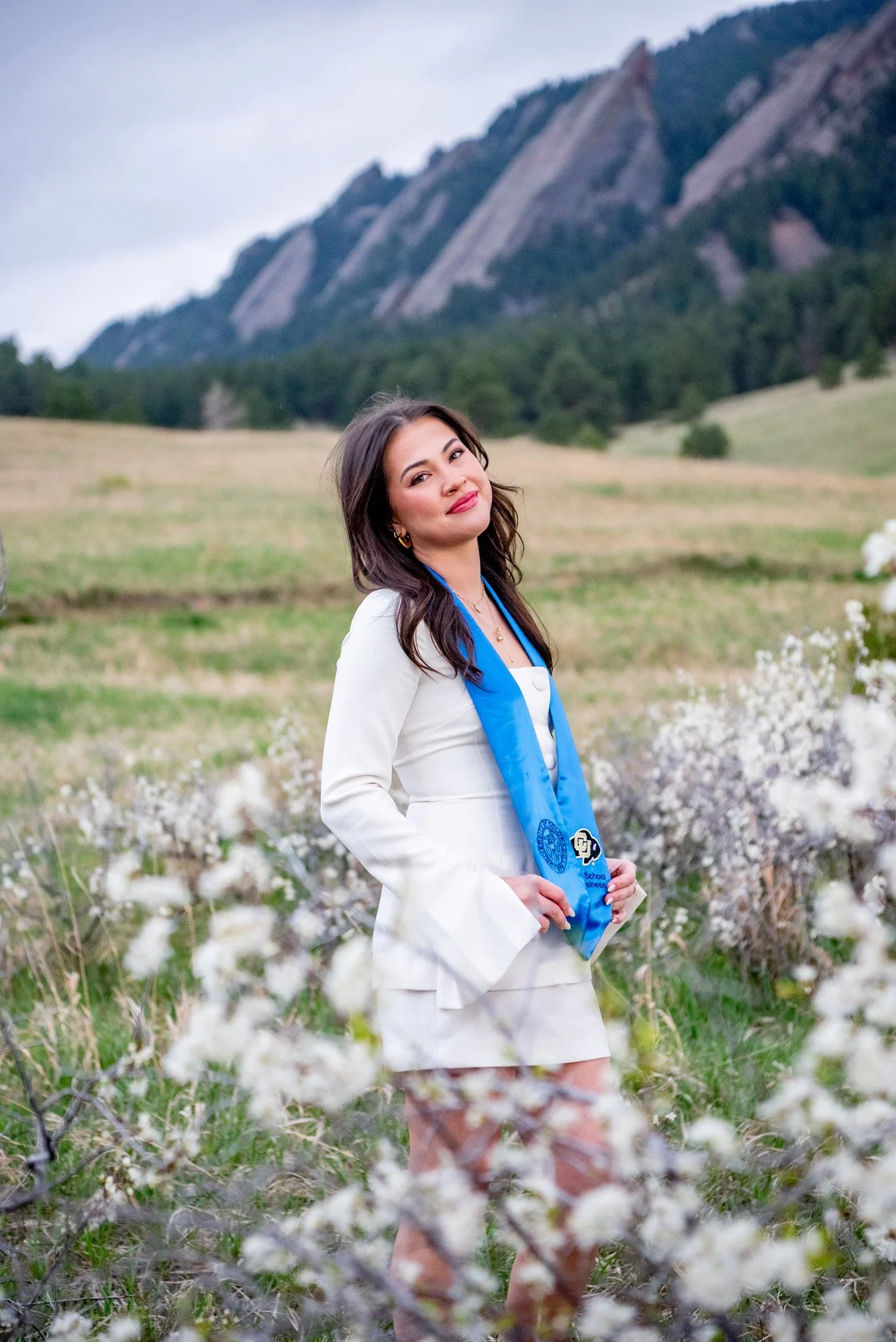 A young woman in a white suit with a blue stole standing in a field of white flowering bushes, mountains in the background.